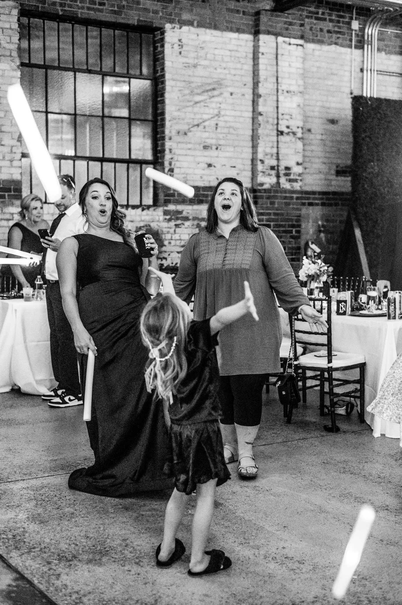 People celebrating at a social event in a rustic indoor setting with exposed brick walls, tables with drinks, and decorations, including a young girl dancing in front of two women who appear surprised and happy.