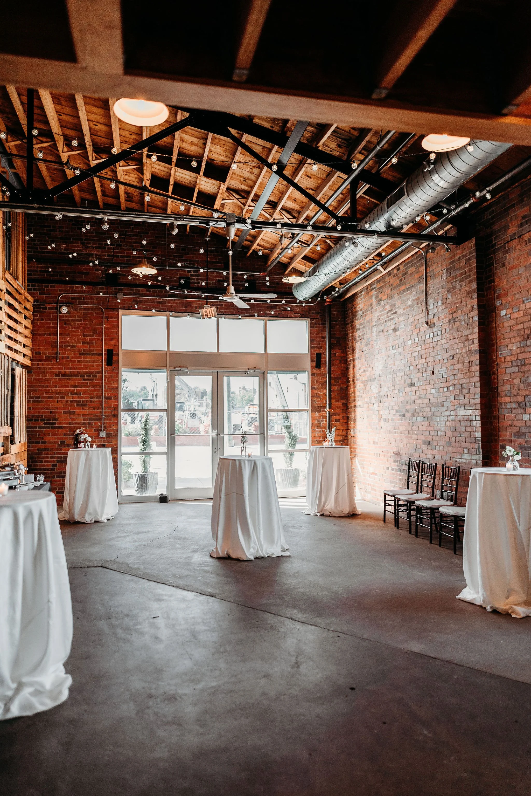 Empty event space with exposed brick walls, wooden ceiling beams, and white high-top tables with draped cloth, decorated with small flower arrangements, and black chairs along the side.