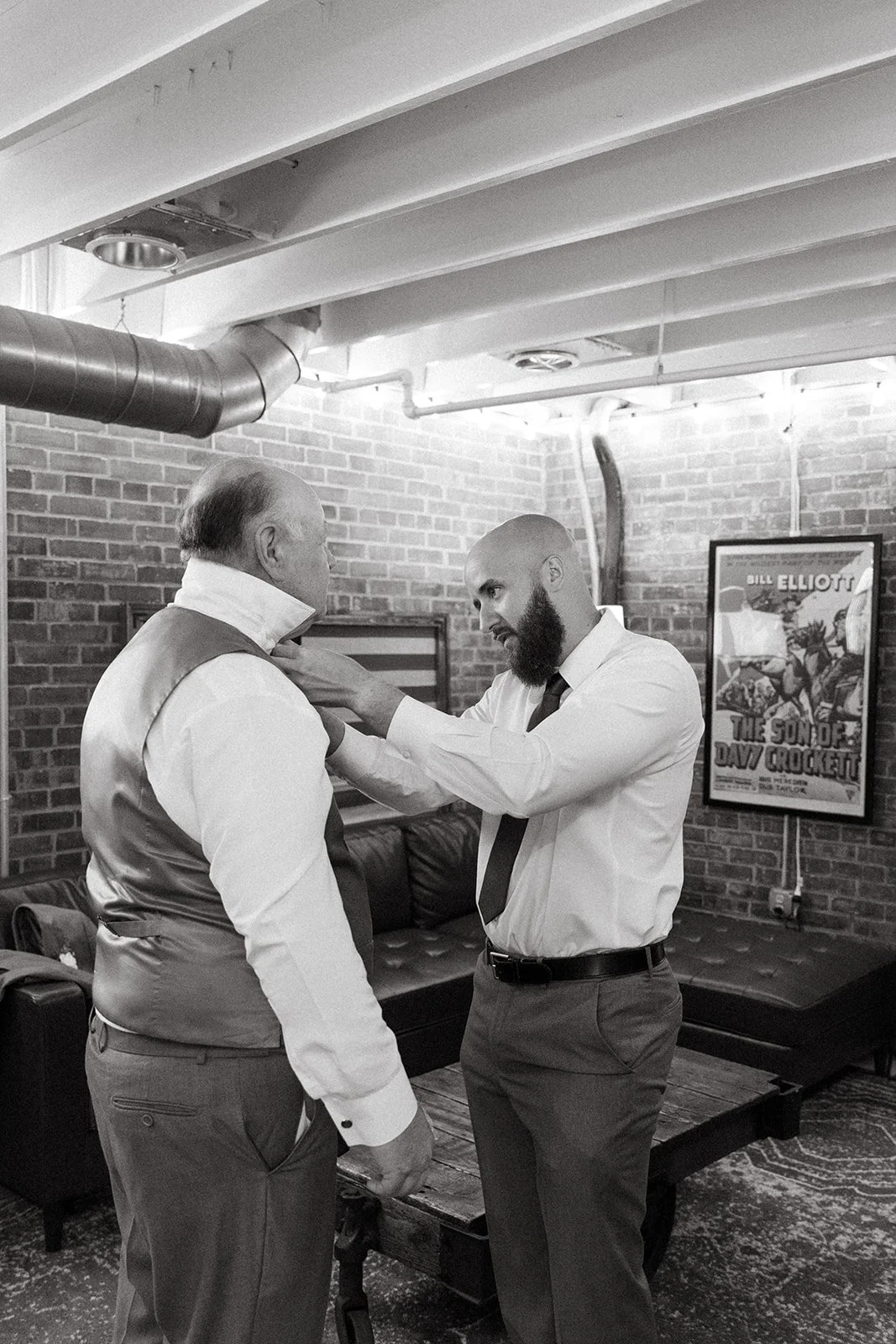 A man with a bald head and beard helping an older man adjust his collar in a room with exposed brick walls, a leather couch, and framed posters on the wall.