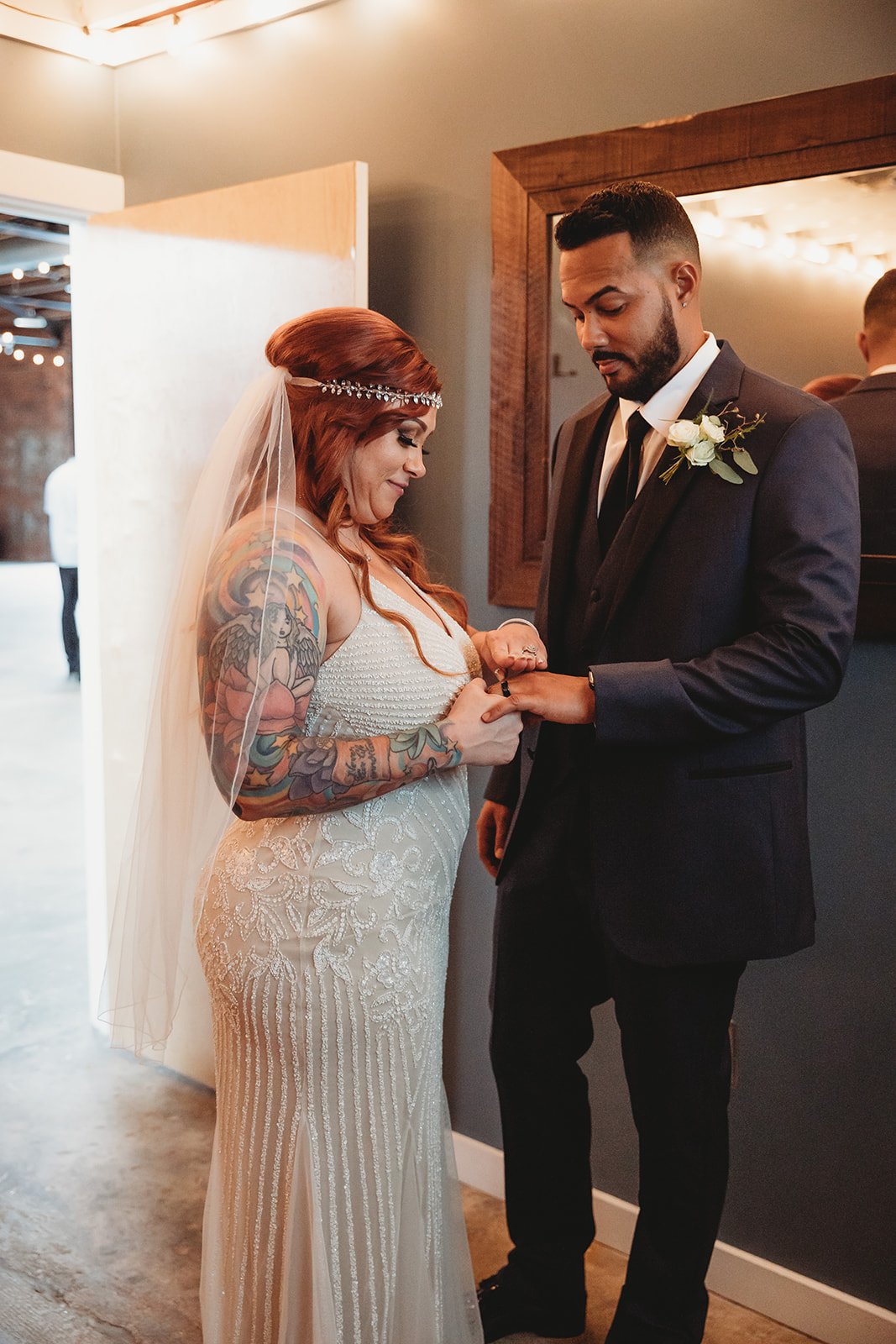 A bride with red hair, tattoos, and a veil is exchanging rings with a groom in a dark suit, during their wedding ceremony indoors.
