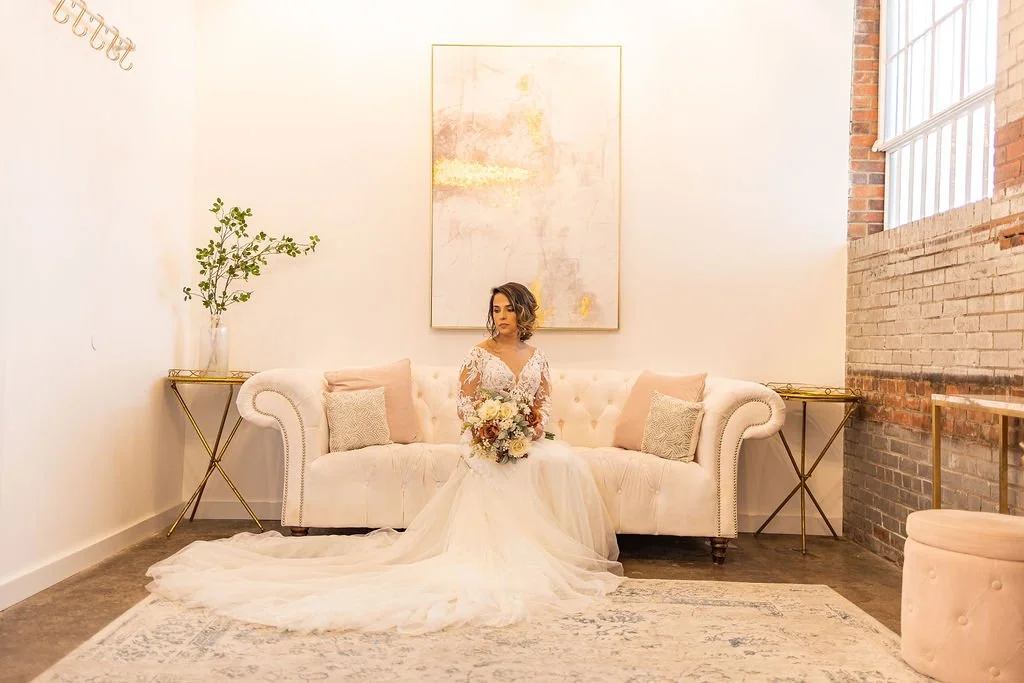 A bride in a white wedding dress sitting on a cream-colored sofa, holding a bouquet of flowers, in a modernas room with white walls, a brick accent wall, a large abstract painting, and a rug.