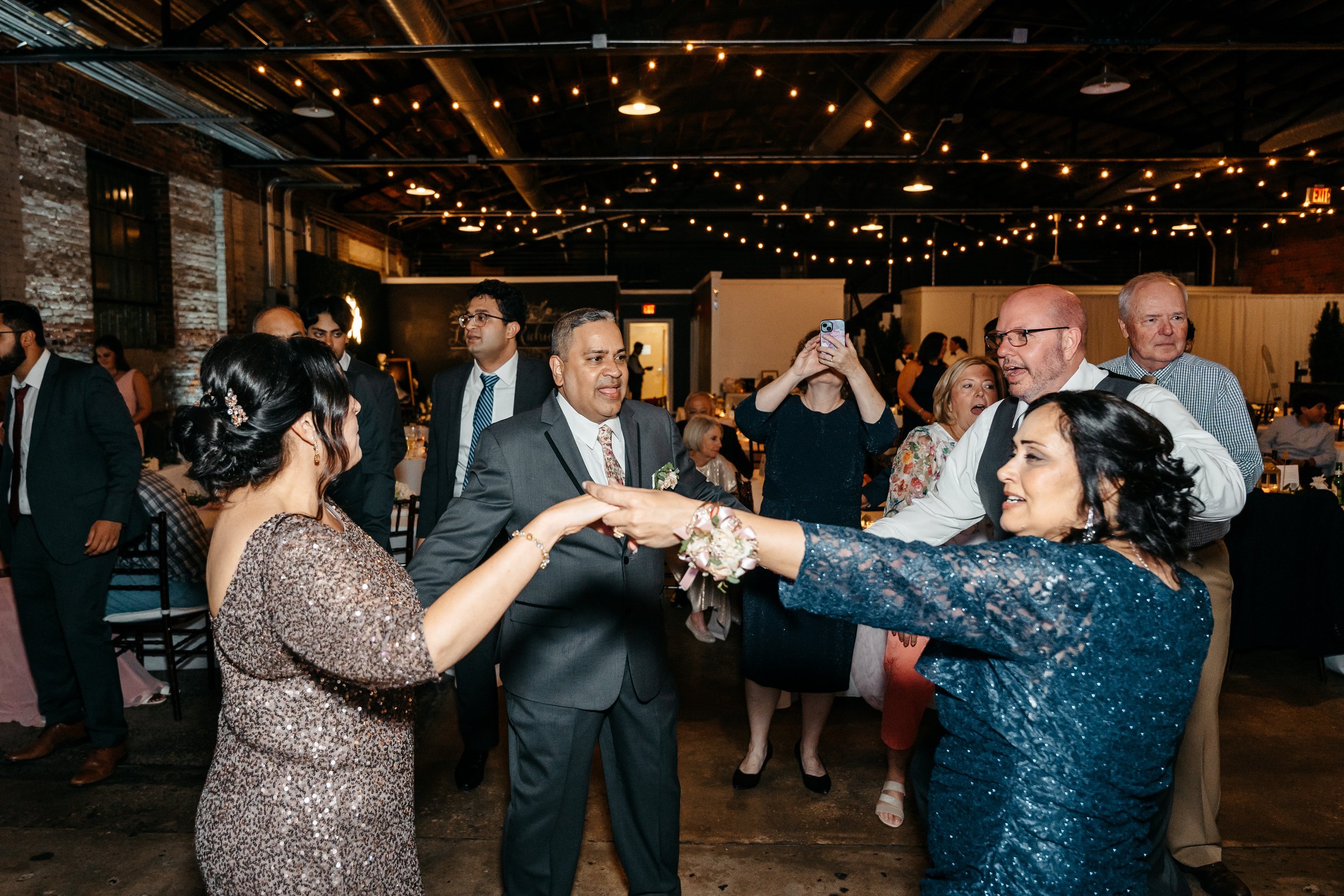 People dancing and socializing at a wedding reception in a decorated venue with string lights overhead.