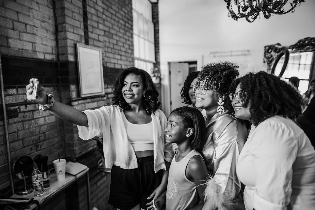 Woman taking a selfie with four women and a young girl in a room with brick wall and large windows.