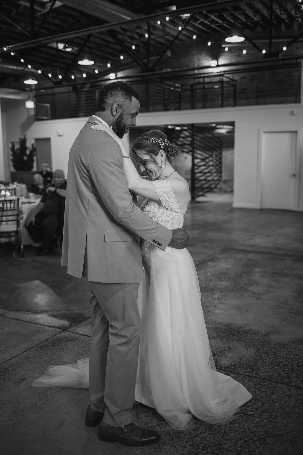 A bride and groom sharing a dance at their wedding reception, smiling and holding each other closely inside a reception hall with string lights overhead.