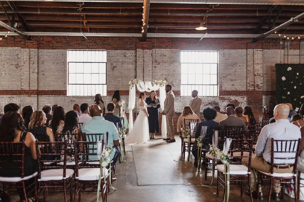 Indoor wedding ceremony in an industrial-style venue with brick walls and large windows, with a bride and groom standing before officiant, surrounded by seated guests.