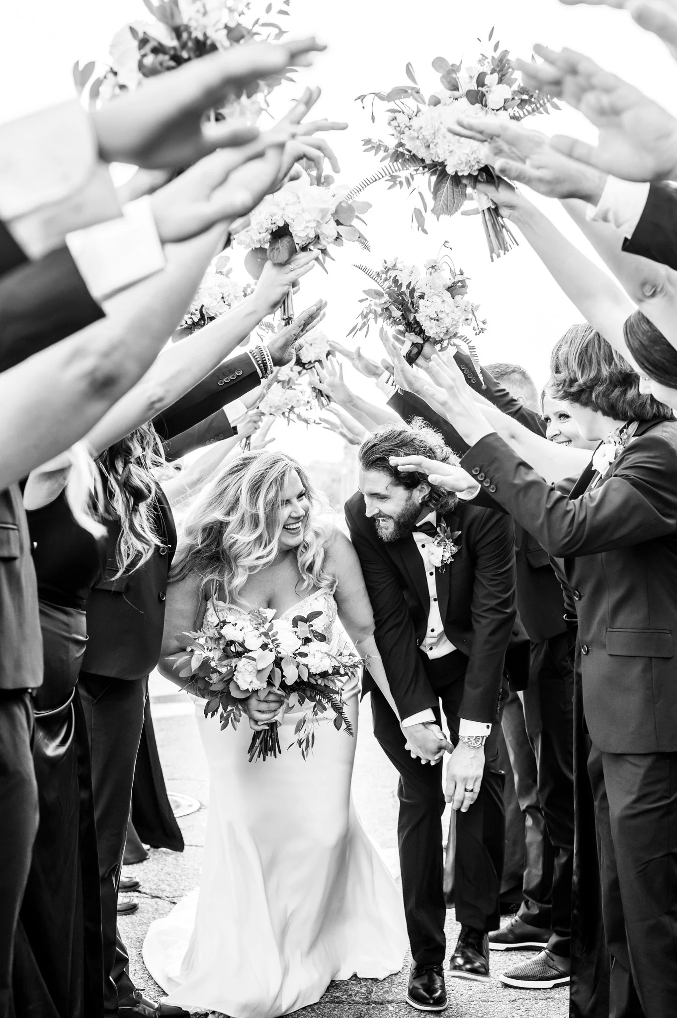 Black and white photo of a bride and groom surrounded by their wedding party, holding bouquets over their heads during a celebratory moment.