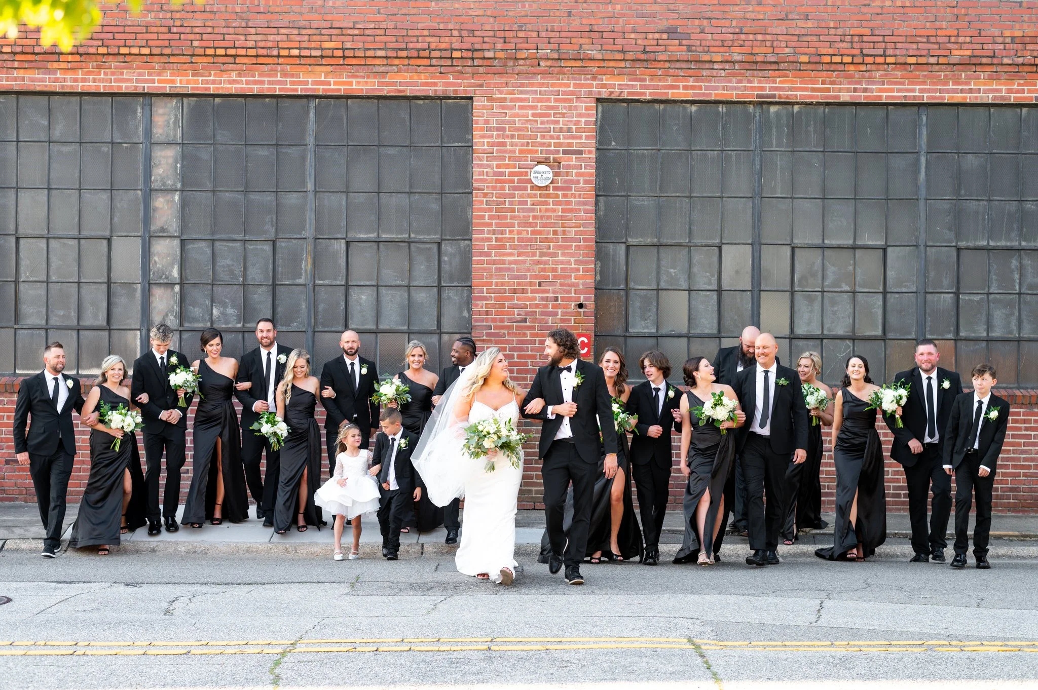 A wedding party walking down a city street in front of a brick building, with bridesmaids in black dresses holding white bouquets, and groomsmen in black suits. The bride and groom are in the center, holding hands and smiling.