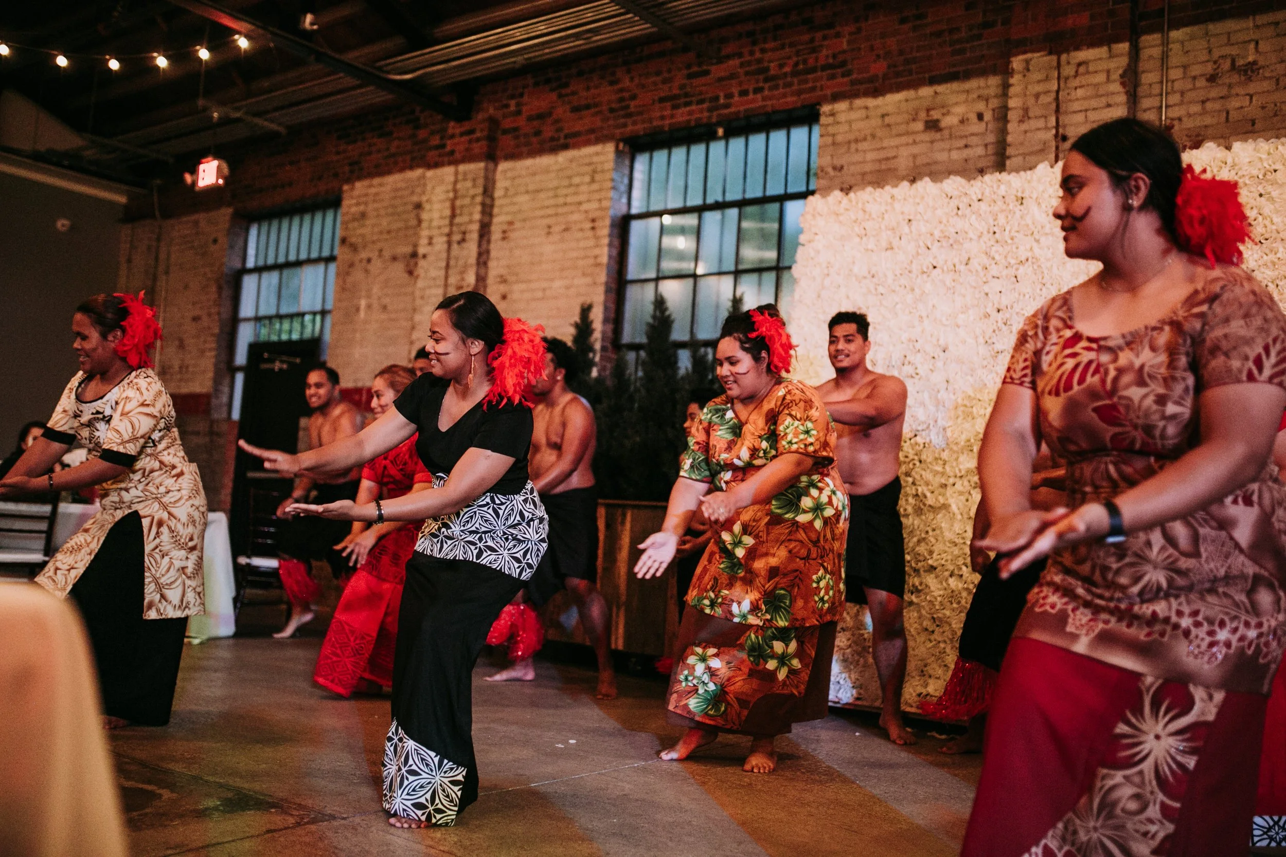 A group of people dancing in traditional Polynesian attire indoors with brick walls and large windows, with women wearing floral dresses and men shirtless.