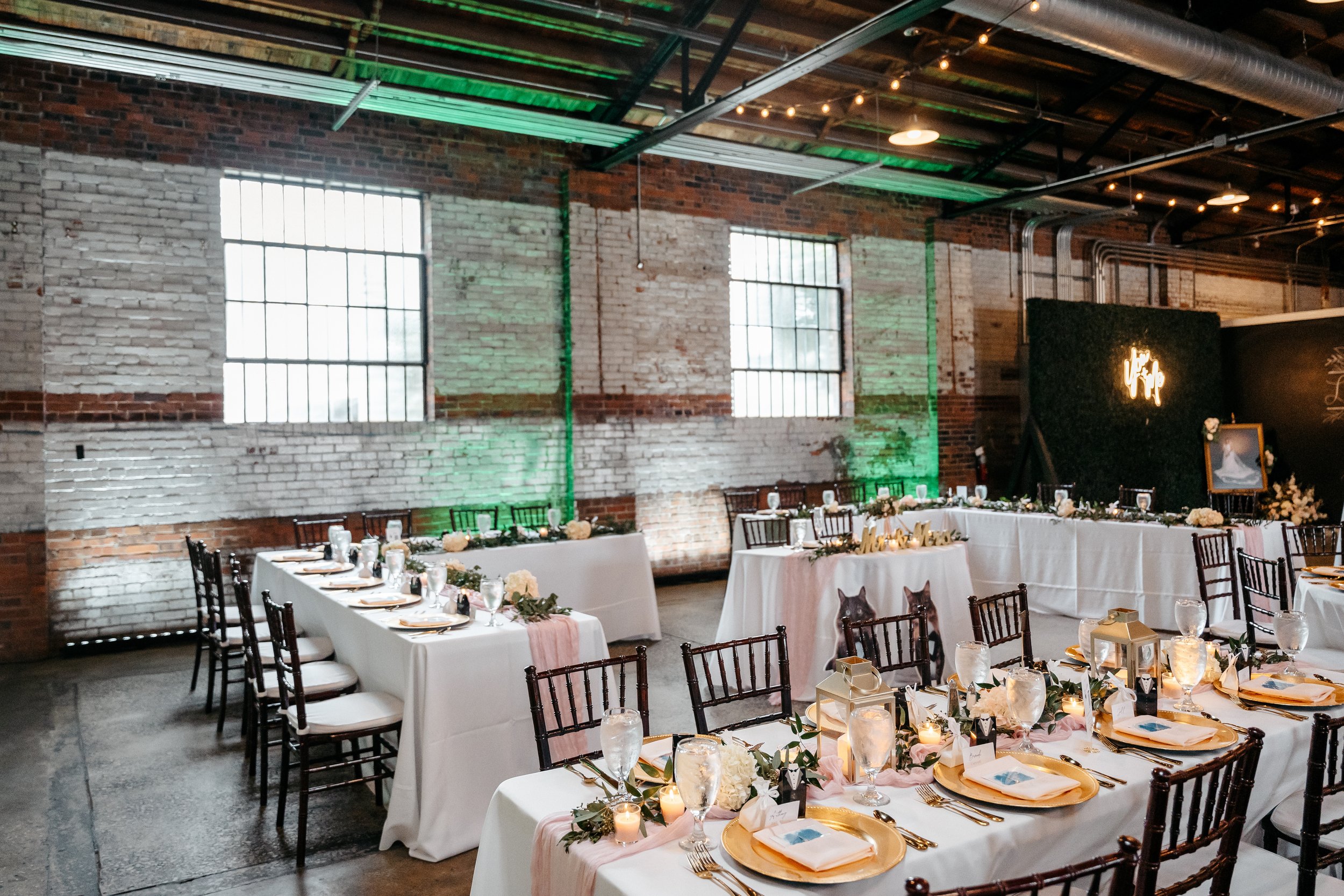 Wedding reception setup with long tables covered in white tablecloths, decorated with candles, flowers, and gold chargers, in a rustic brick and concrete industrial venue.