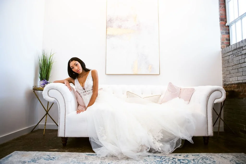 A woman in a white dress sitting on a white couch in a bright, modern room with a large window and brick wall.