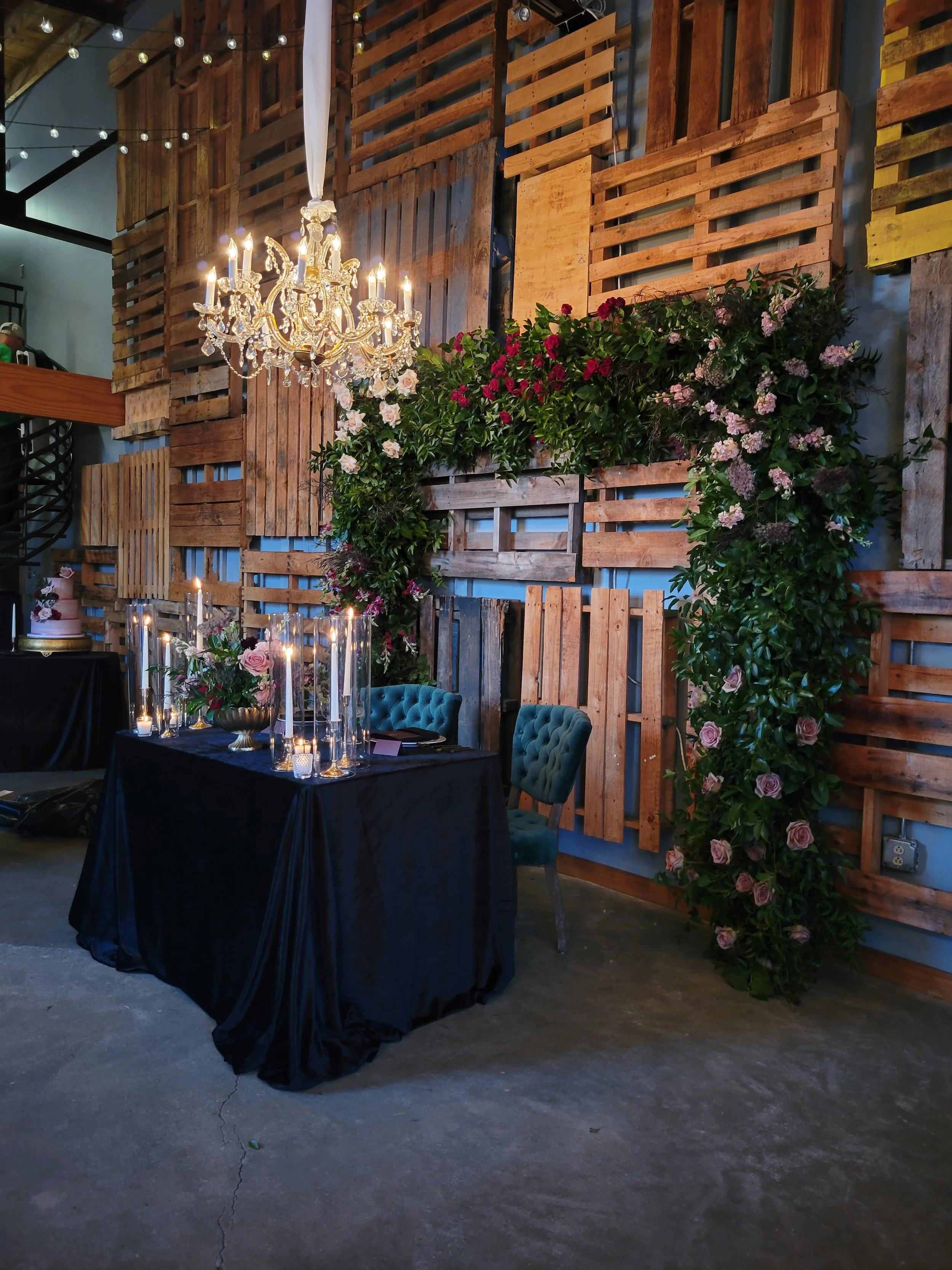 Elegant event setup featuring a black tablecloth-covered table with candles and floral arrangements, a vintage blue upholstered chair, a rustic wooden pallet backdrop with pink and white flowers, and a chandelier hanging from the ceiling.