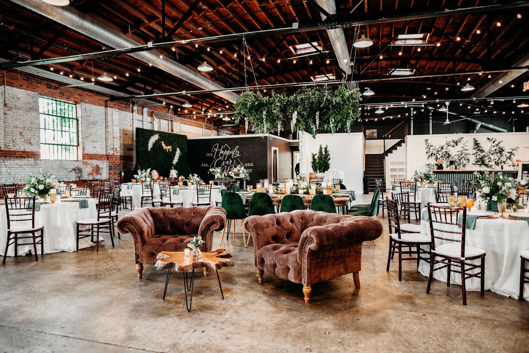 Wedding reception setup in a rustic industrial venue with round tables, white tablecloths, green chairs, a hanging greenery installation, a lounge area with vintage sofas, and string lights overhead. at Studio 215 a modern venue in Fayetteville