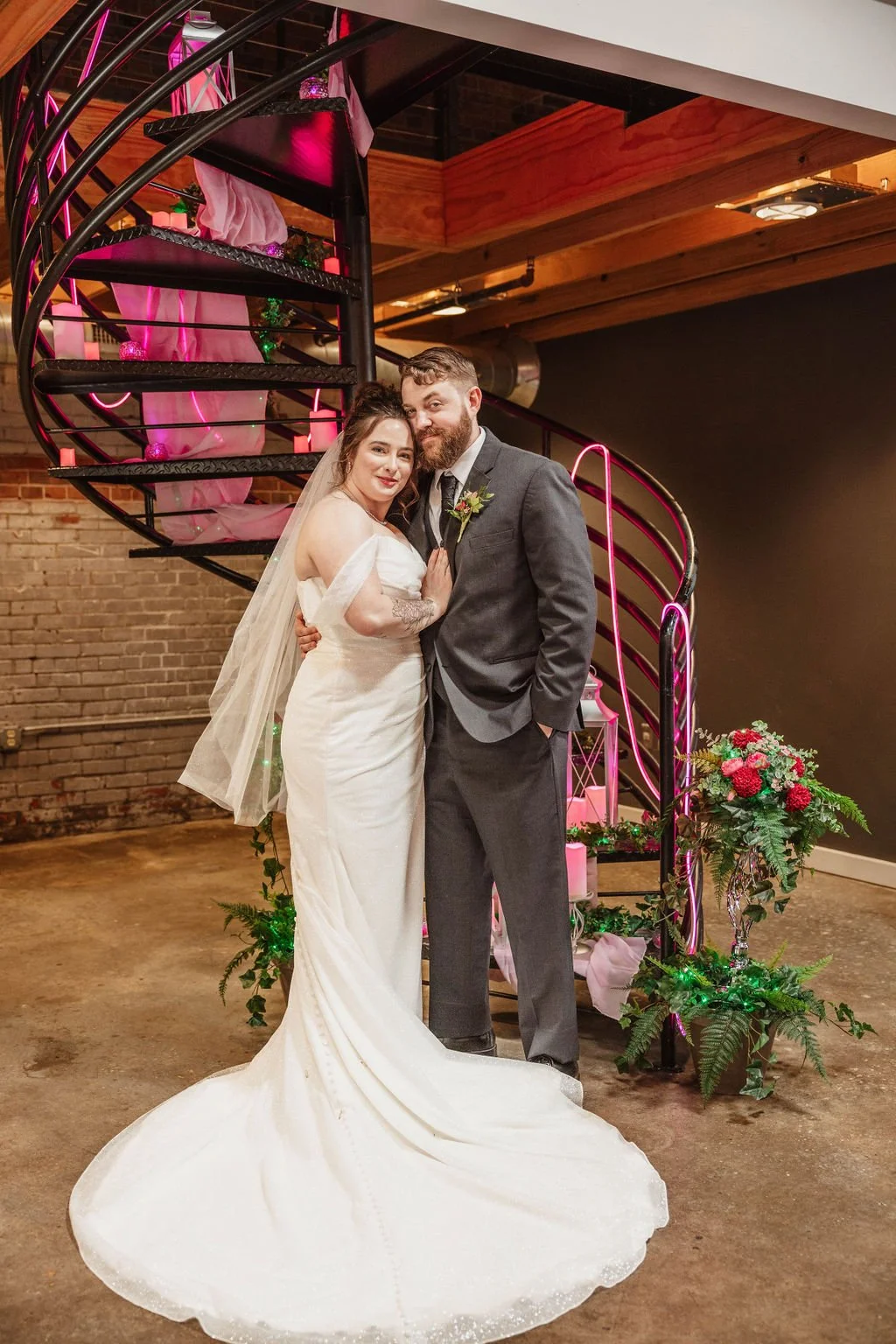 A bride and groom standing together on their wedding day, posing in front of a spiral staircase decorated with pink and green lights and flowers, inside a rustic venue with exposed brick wall and wooden ceiling.