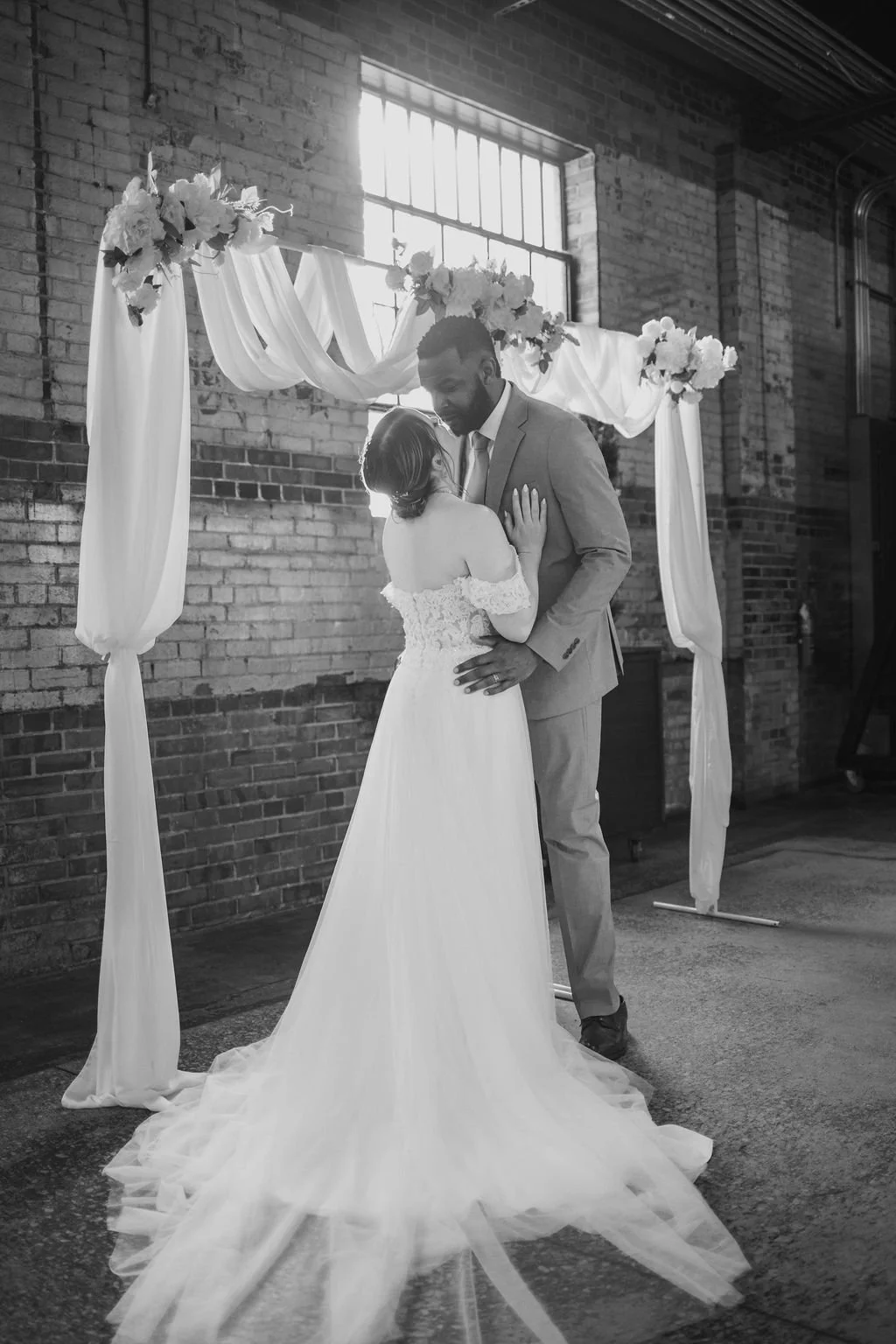 A couple in wedding attire sharing a dance under a floral and fabric wedding arch, with a brick wall and window in the background.
