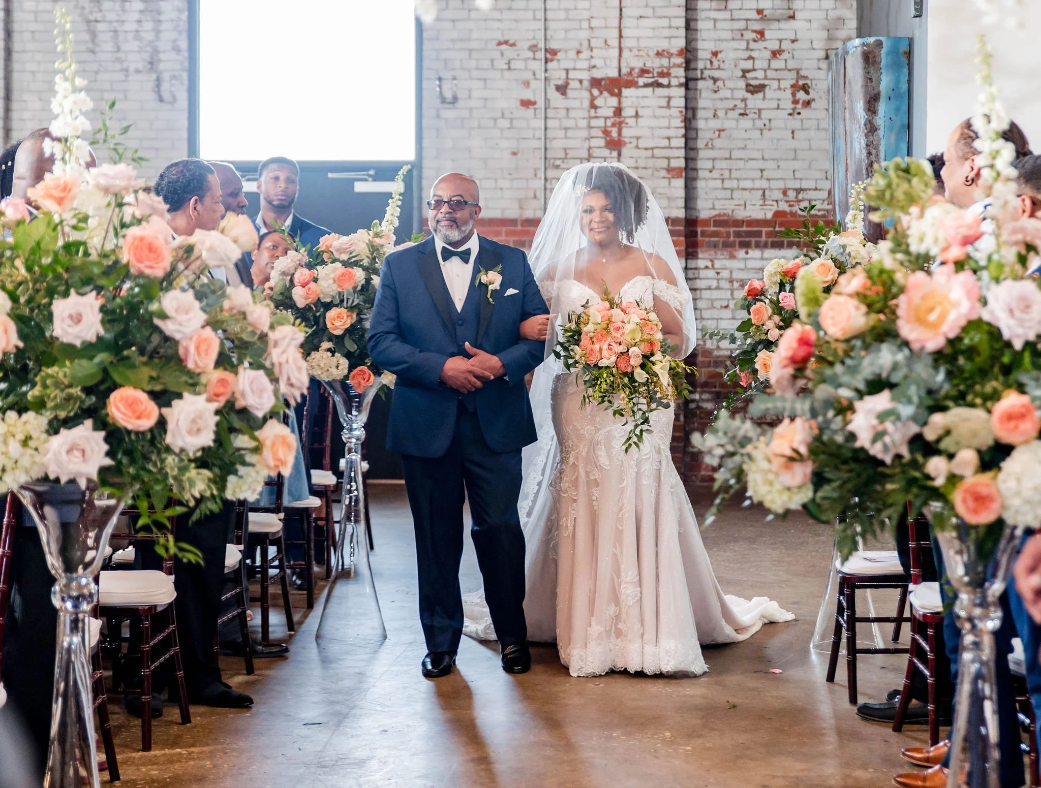 A bride and an older man walking down the aisle during a wedding ceremony in a rustic venue with brick walls and large windows, surrounded by floral arrangements of pink, white, and peach flowers.