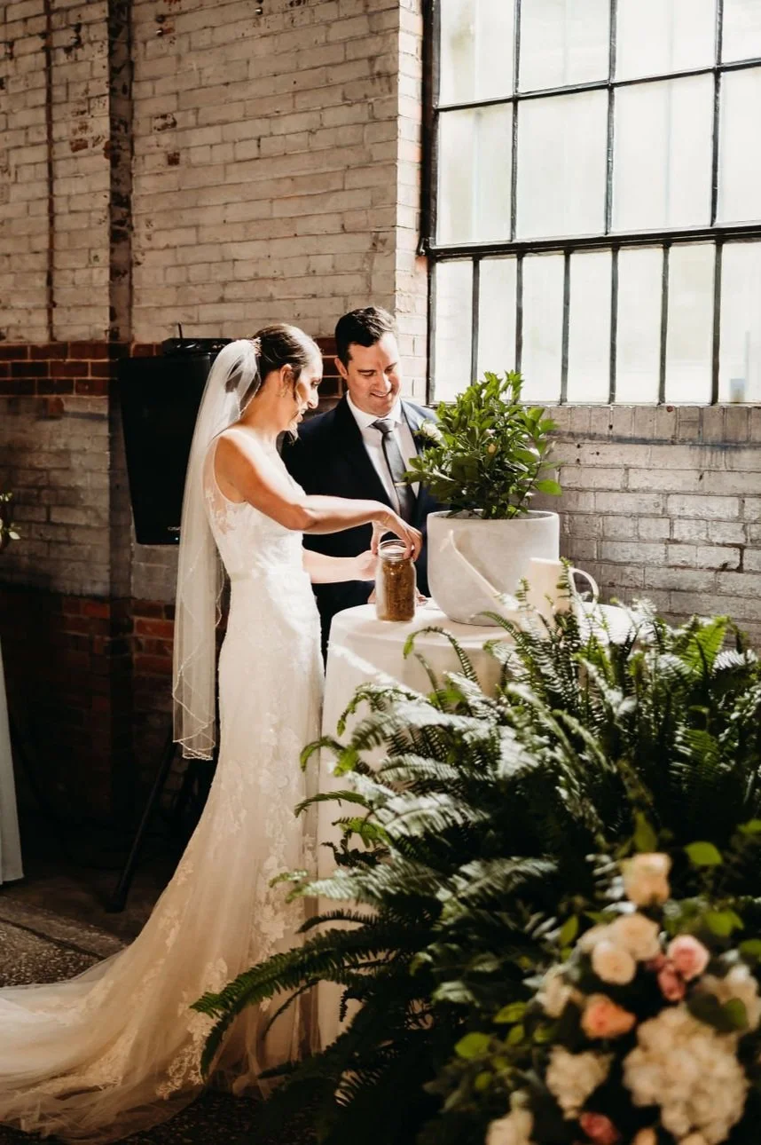 A bride and groom planting a small tree or shrub in a white pot during their wedding ceremony inside a rustic venue with exposed brick walls and large industrial-style windows, decorated with greenery and flowers.
