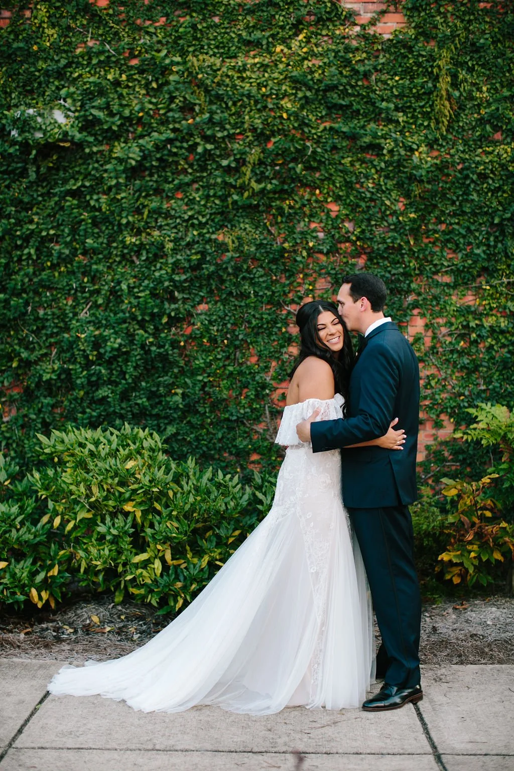 A newlywed couple embracing outdoors in front of a brick wall covered with green ivy, the bride in a white wedding gown smiling and the groom in a navy suit.