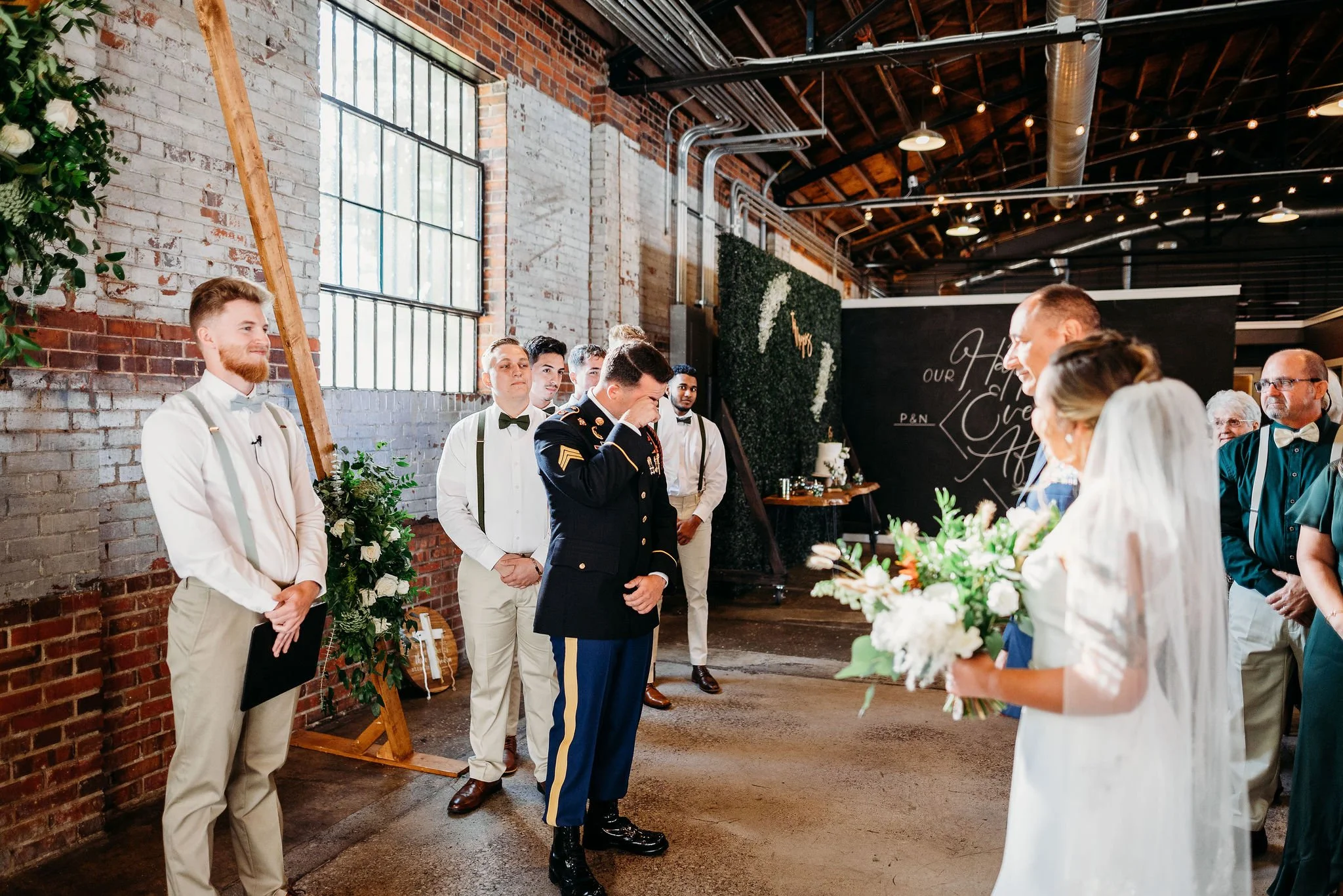 A wedding ceremony in an industrial-style venue with brick walls, large windows, and hanging lights. The bride is holding a bouquet, the groom is in a tuxedo, and several men in formal attire are standing nearby, including a man in a military uniform