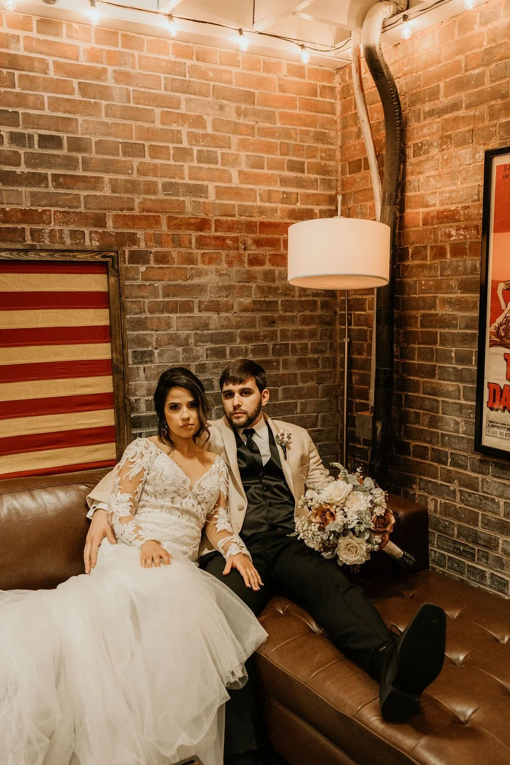 A bride and groom sitting together on a brown leather couch in a rustic, industrial-style setting with a brick wall background. The bride wears a white lace wedding dress, and the groom is dressed in a beige blazer, black shirt, and tie, holding a large bouquet of flowers.