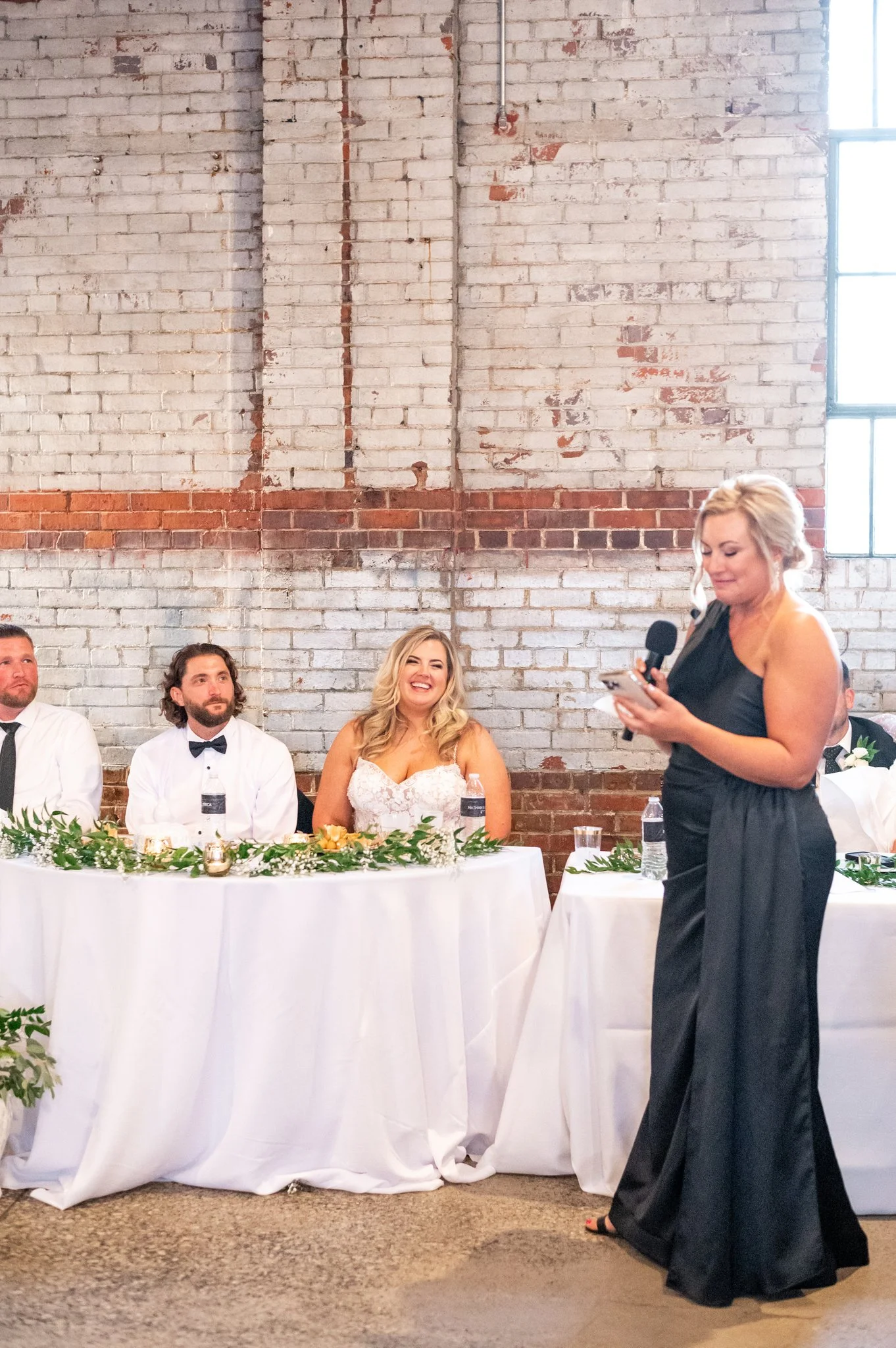 A woman giving a speech at a wedding reception, standing next to a long table with seated guests. The bride, in a white wedding dress, is smiling and sitting between two men in white shirts with black ties. The setting has an industrial brick wall ba
