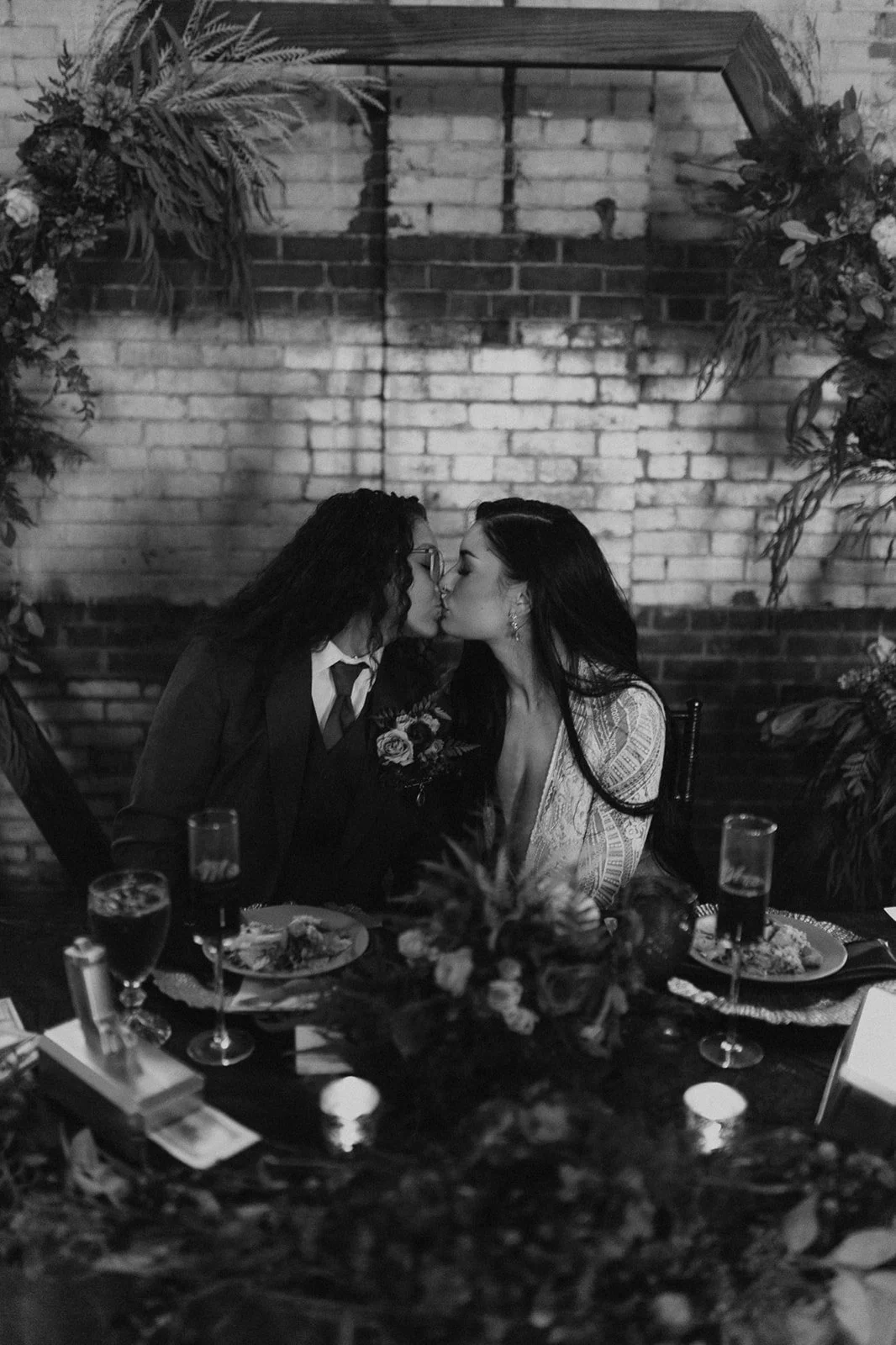 Two women sharing a kiss at a wedding or romantic dinner, sitting at a decorated table with flowers and drinks, in front of a brick wall backdrop.