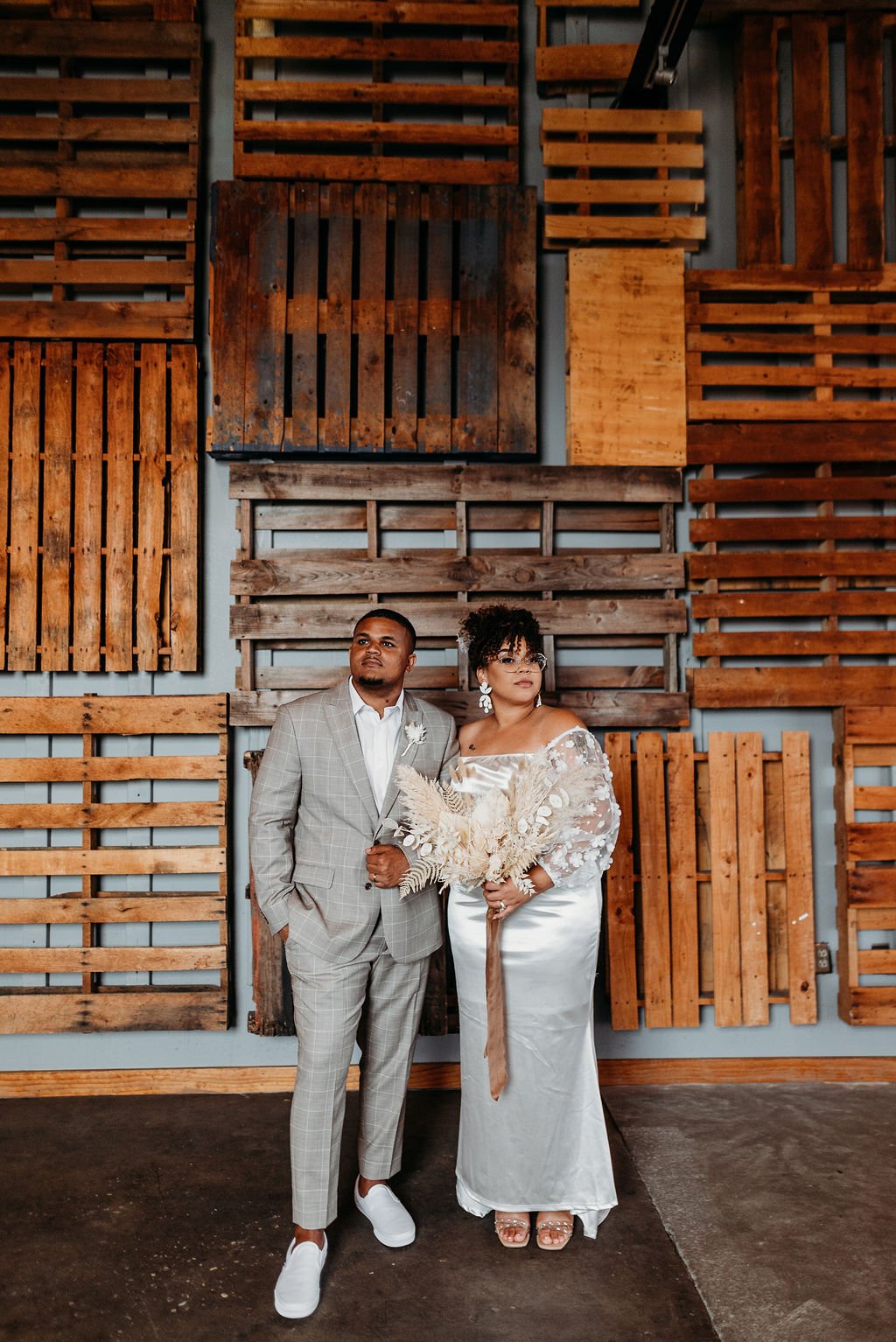 A couple dressed in wedding attire standing in front of a rustic wooden wall with various wooden panels as decor.