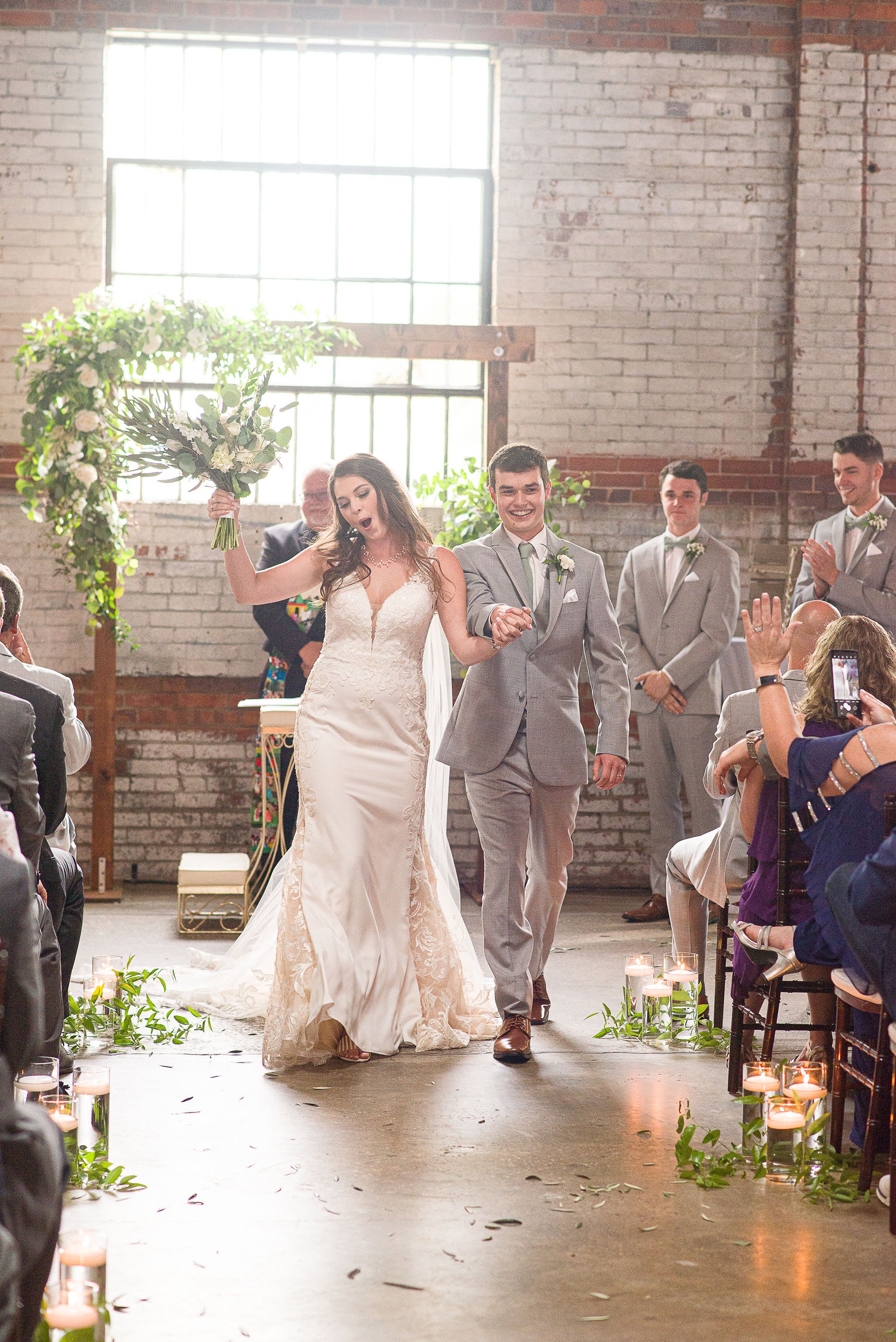 A newly married couple walking down the aisle at their wedding ceremony, with guests on both sides and a floral arch in the background, inside a rustic industrial venue with brick walls.