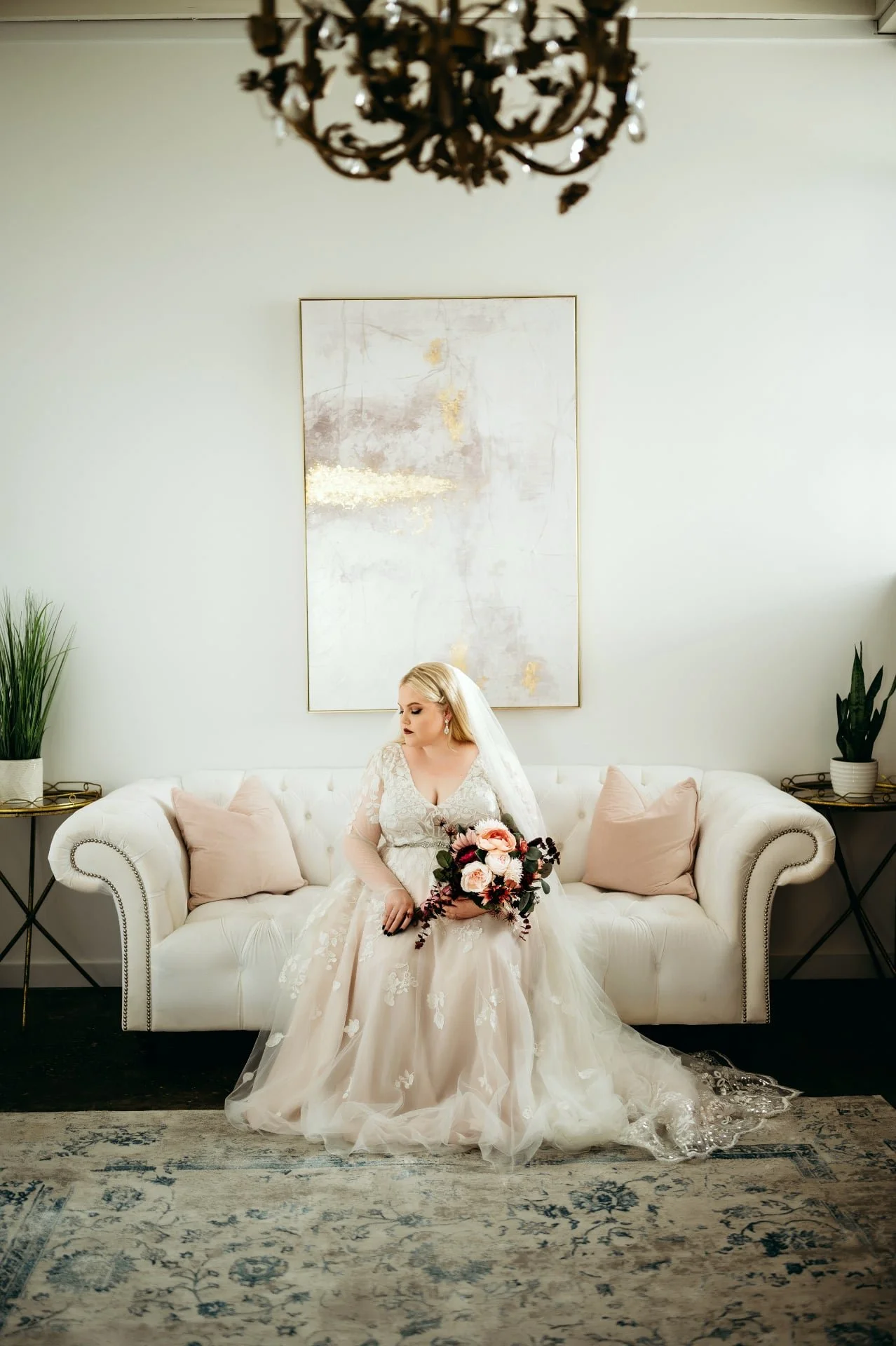 A woman in a wedding dress sitting on a white tufted sofa holding a bouquet of pink roses and greenery, with pale pink pillows on either side, in a room with a painting on the wall, plants on side tables, and a chandelier overhead.