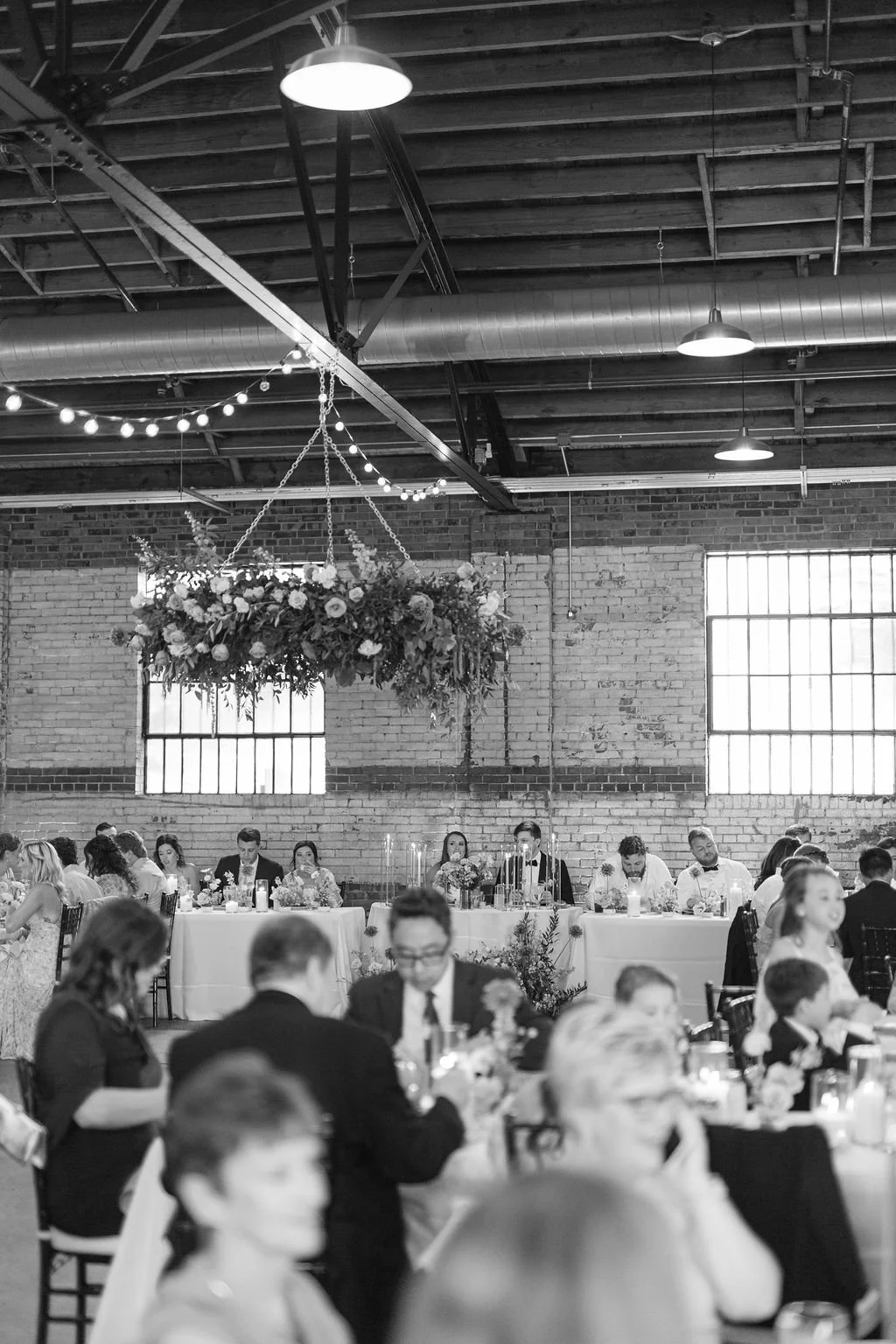 Indoor wedding reception with long head table, floral arrangements, hanging floral chandelier, string lights, and brick walls with large windows.