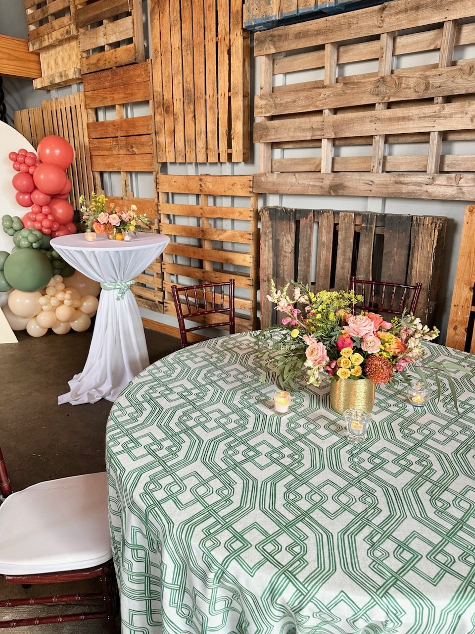 Elegant event setup at Little Studio 215 featuring a green geometric patterned tablecloth, gold floral centerpieces, and a rustic wood pallet backdrop with coral and sage balloon installations.