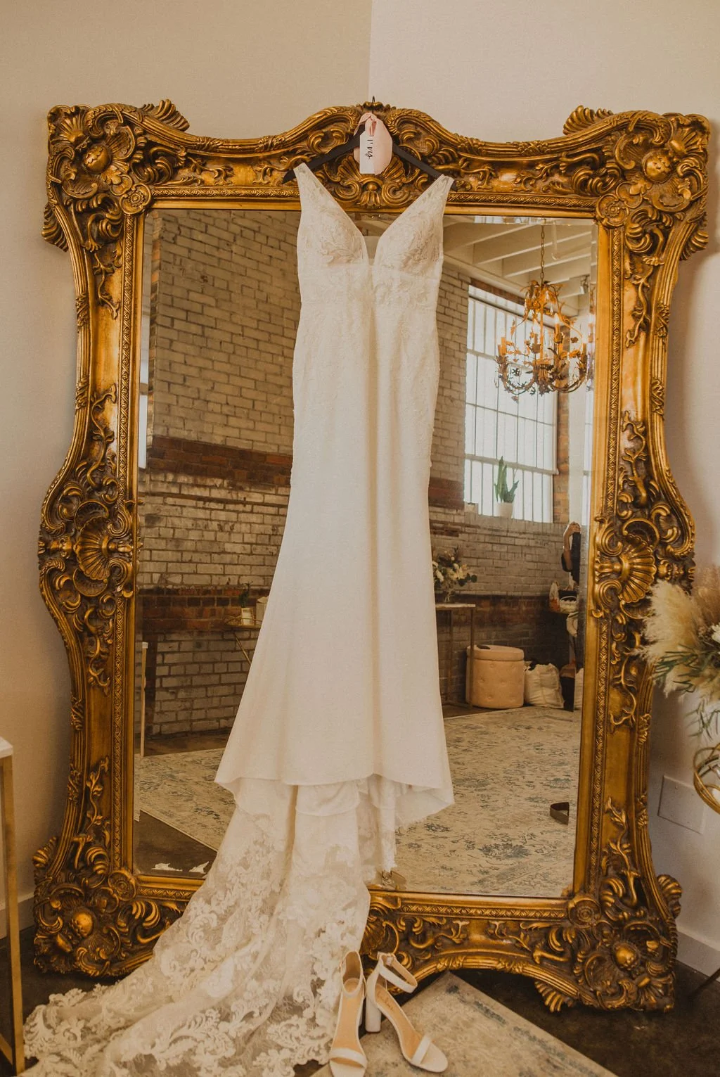 Wedding dress hanging in front of a large ornate gold mirror with a train laying on the floor, white heels underneath, in a room with brick walls, large windows, and a chandelier.