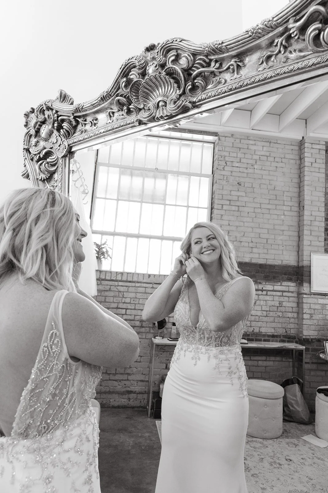 Two women in wedding dresses smiling and adjusting earrings in front of a large ornate mirror in a loft style room with large windows and brick walls.