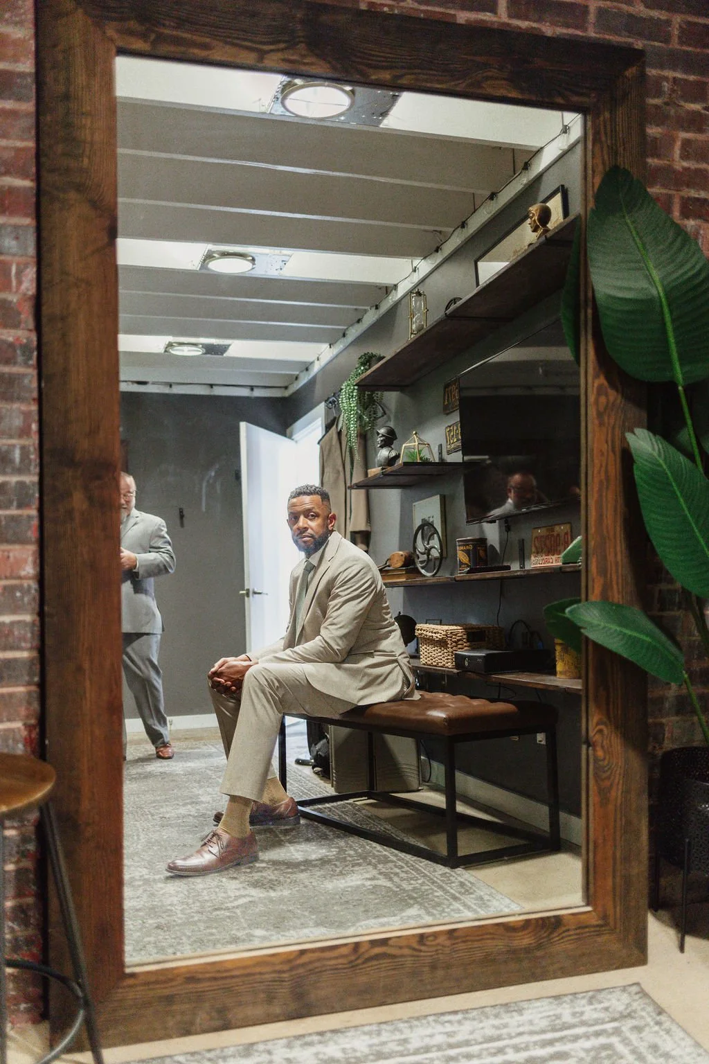 Reflection of a man in a mirror sitting on a bench in an industrial-style room with shelves, plants, and another person in the background.