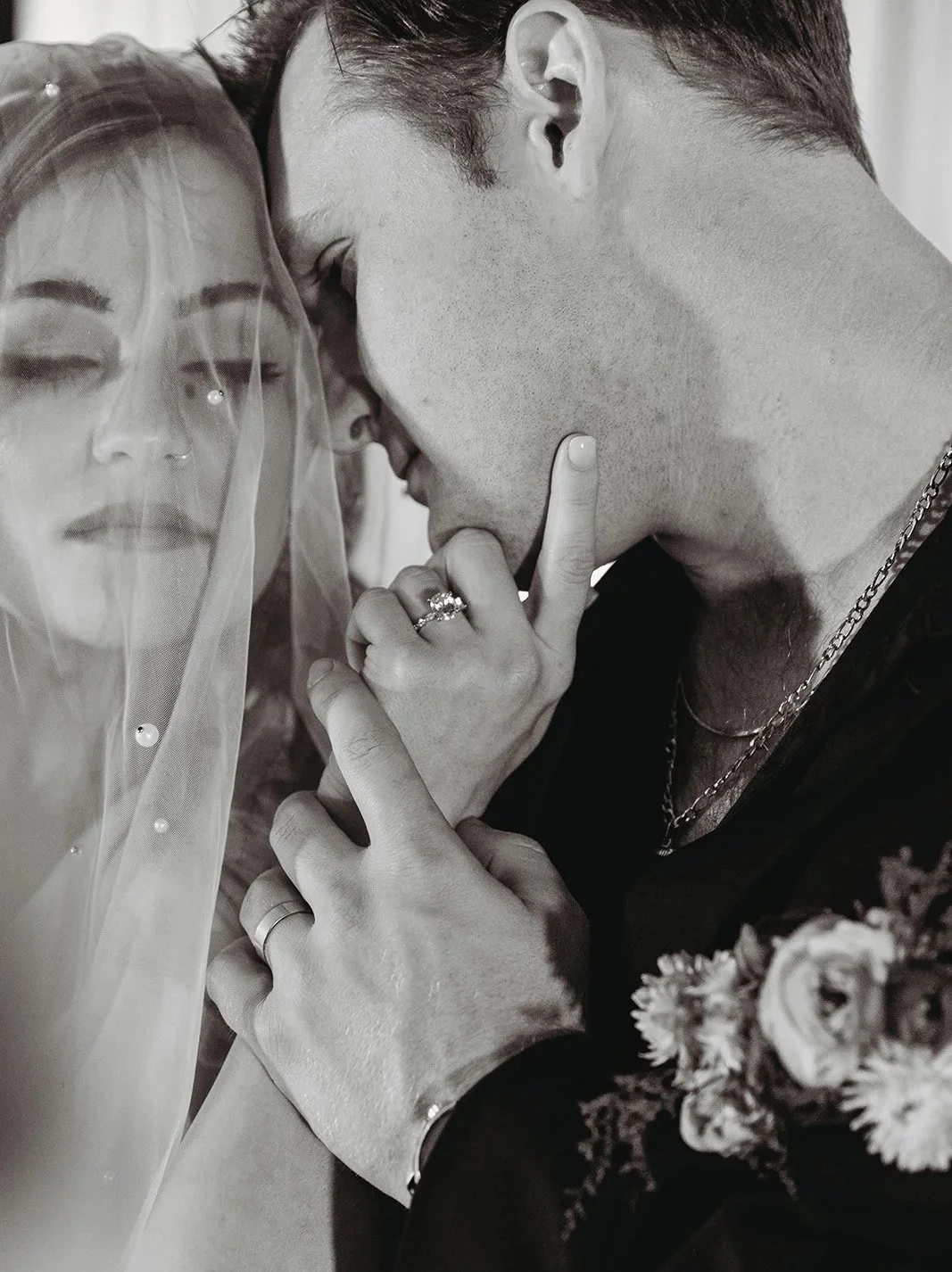 A black-and-white photo of a couple in an intimate moment. The woman is partially covered with a veil and the man is touching her lips with his finger, both with their eyes closed.