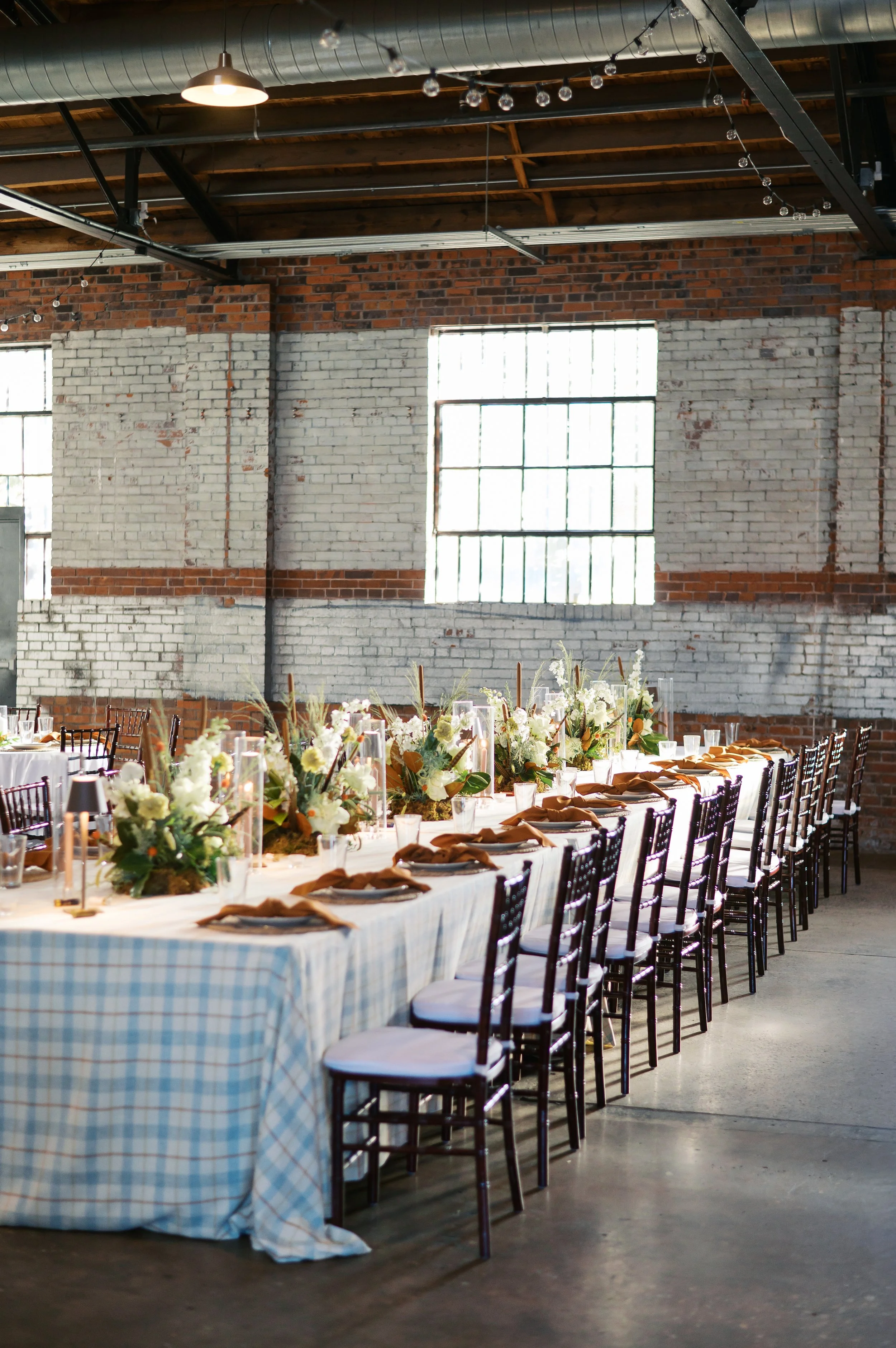 Long banquet table decorated with flowers, candles, and place settings in an industrial-style event space with brick walls and large windows.