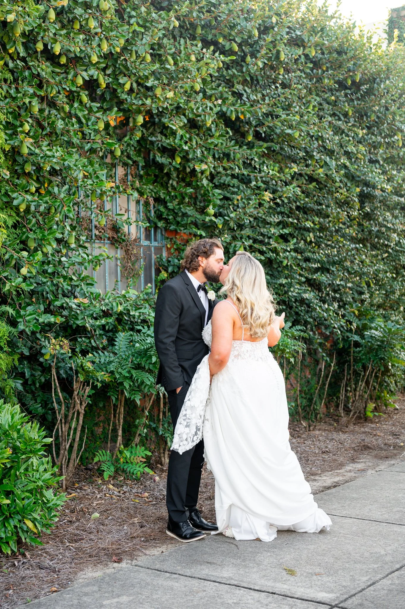 A newlywed couple sharing a kiss outdoors, standing on a sidewalk next to a lush green hedge.