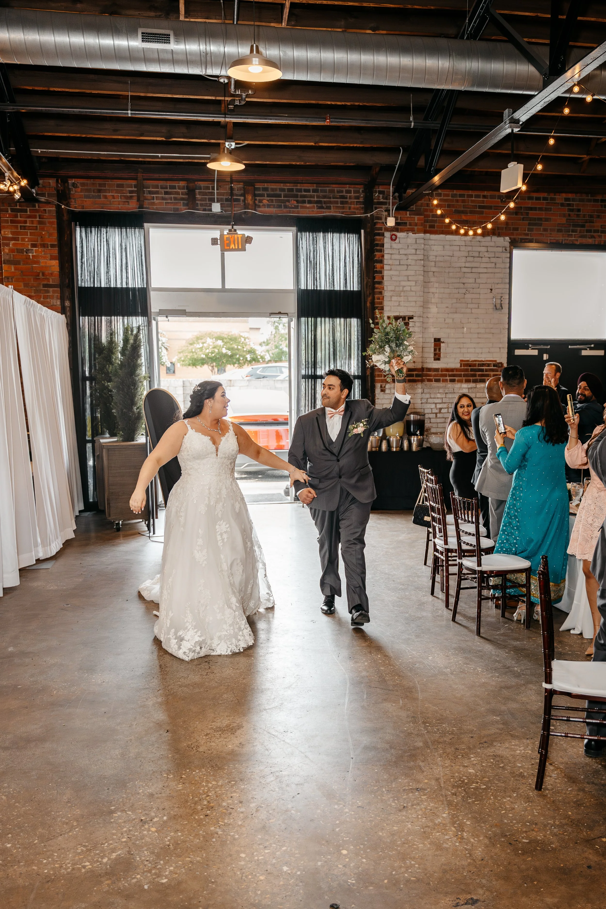 A bride and groom dancing and celebrating at their wedding reception in a decorated industrial-style venue, with guests watching and taking photos.