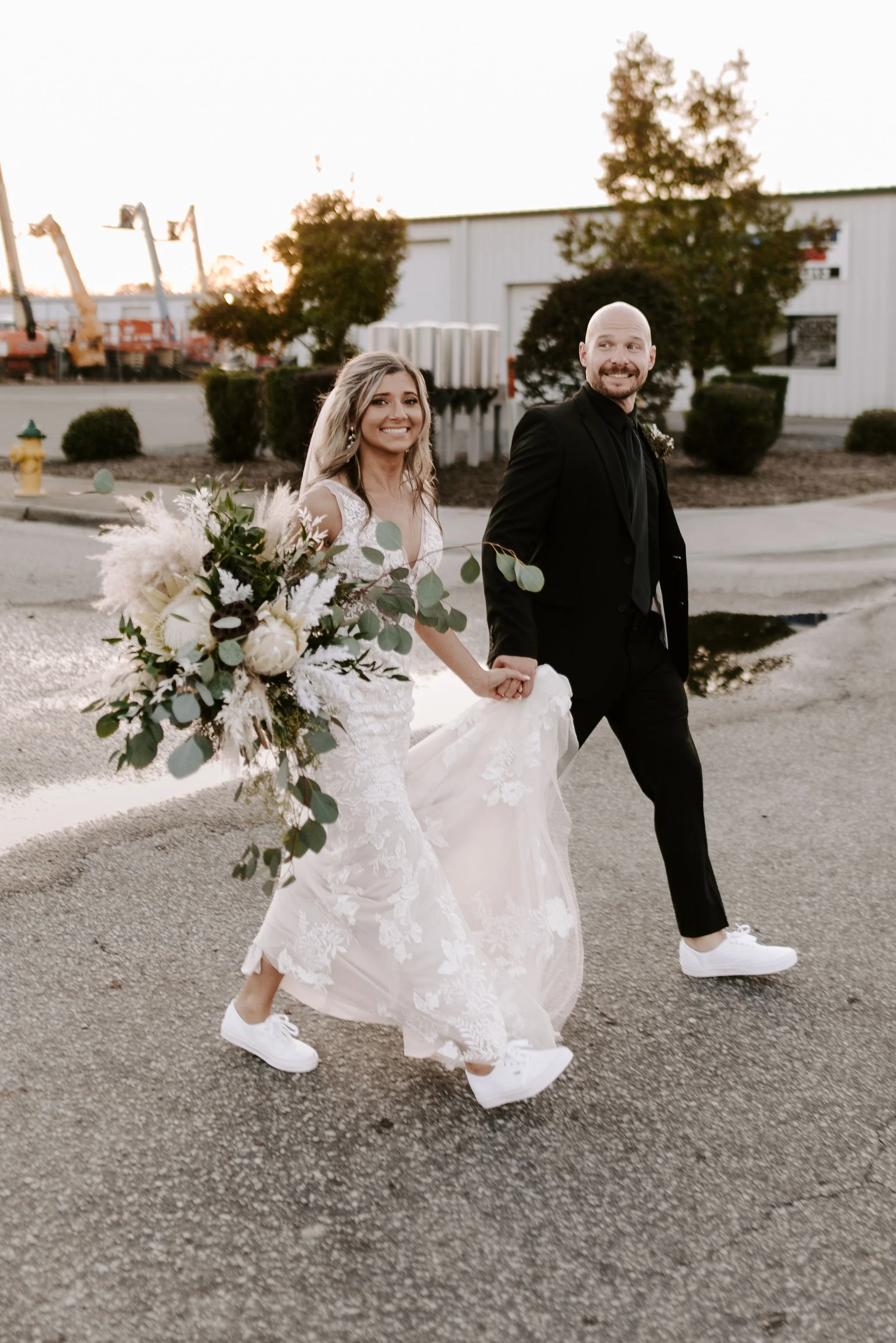 A newlywed couple walking outdoors holding hands, with the bride in a lace wedding dress and the groom in a black suit, both wearing white sneakers, during sunset.