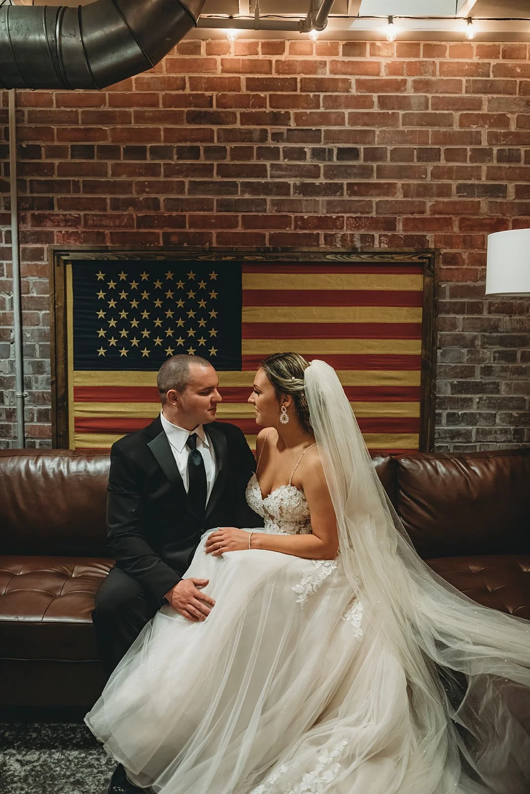 A bride and groom sitting on a leather couch in front of a brick wall with an American flag hanging behind them. The bride is wearing a white wedding gown and veil, and the groom is in a black suit and tie. They are looking into each other's eyes.