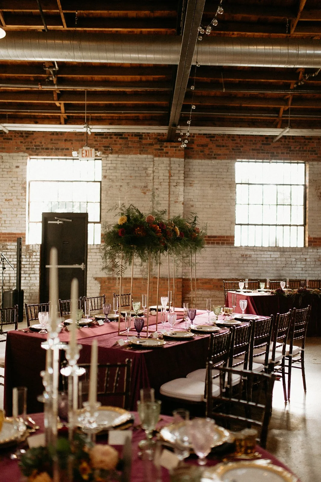 Indoor event space with brick walls and large windows, decorated for a formal dinner with a long table covered in a wine-colored tablecloth, set with plates, silverware, and glasses, and adorned with a tall floral centerpiece with green foliage and c