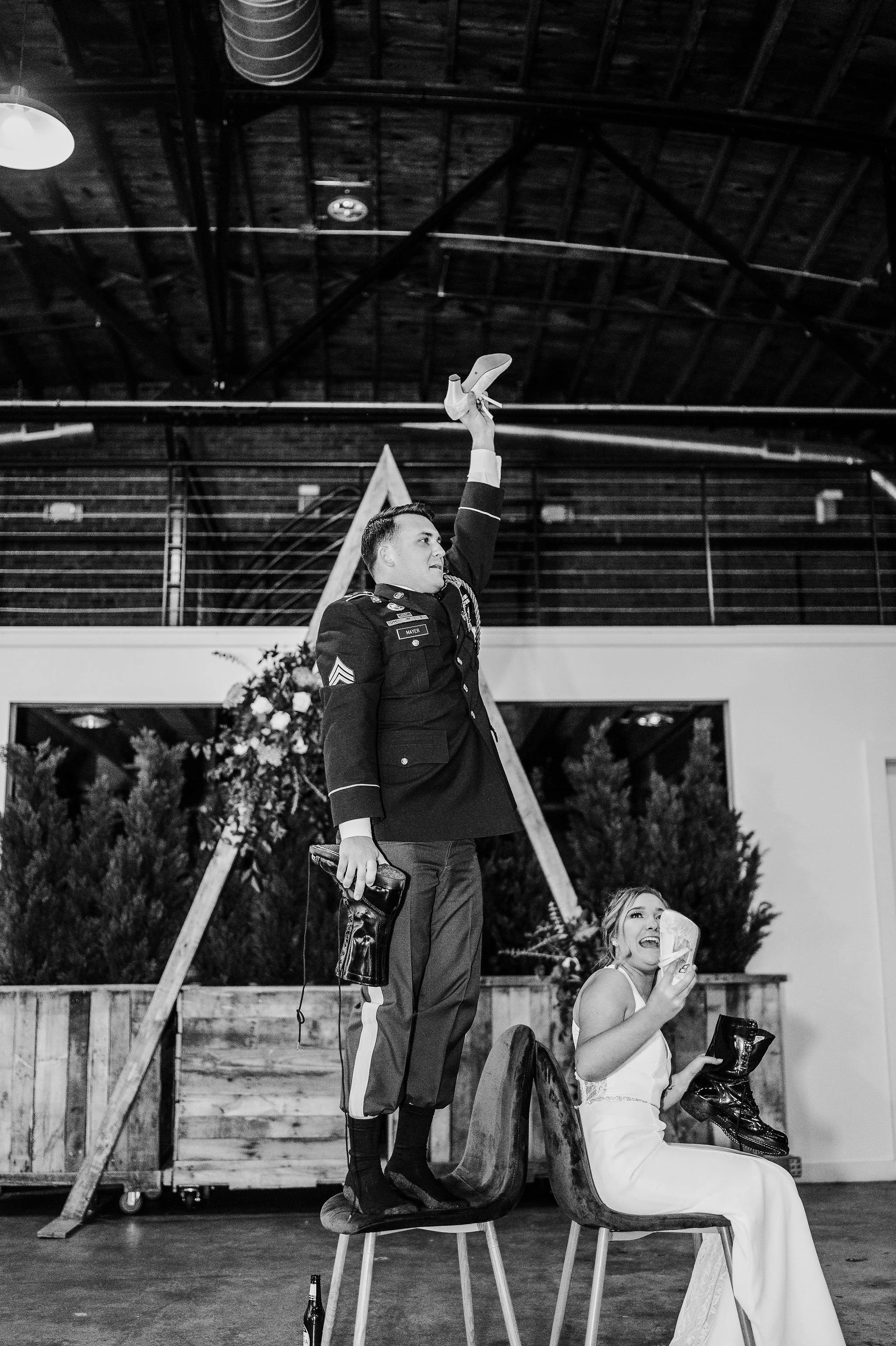 Black and white photo of a man in a military uniform standing on a chair with one arm raised and holding a shoe, and a woman in a white dress sitting on a chair next to him, holding a shoe and a tissue, in an indoor setting with a wooden backdrop and