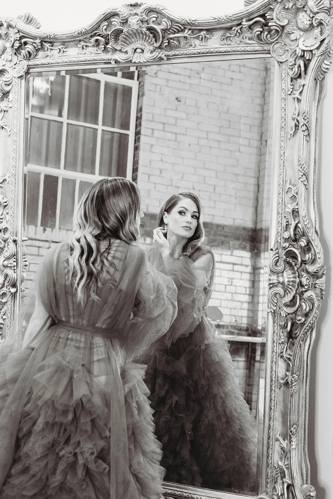 A woman in a ruffled dress looking at herself in a large ornate mirror with an intricate frame, in a room with brick walls.