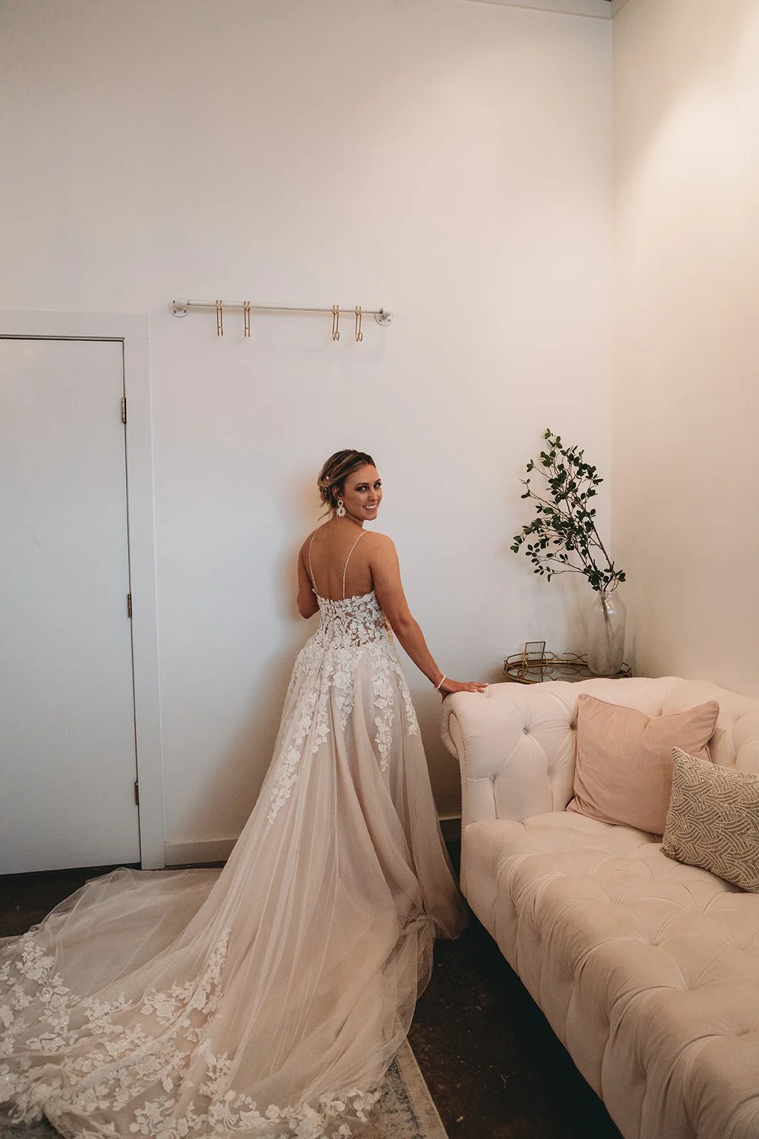 Bridal woman in a wedding dress standing beside a beige tufted sofa with pillows, a side table, and a vase with green foliage in a minimalistic room.