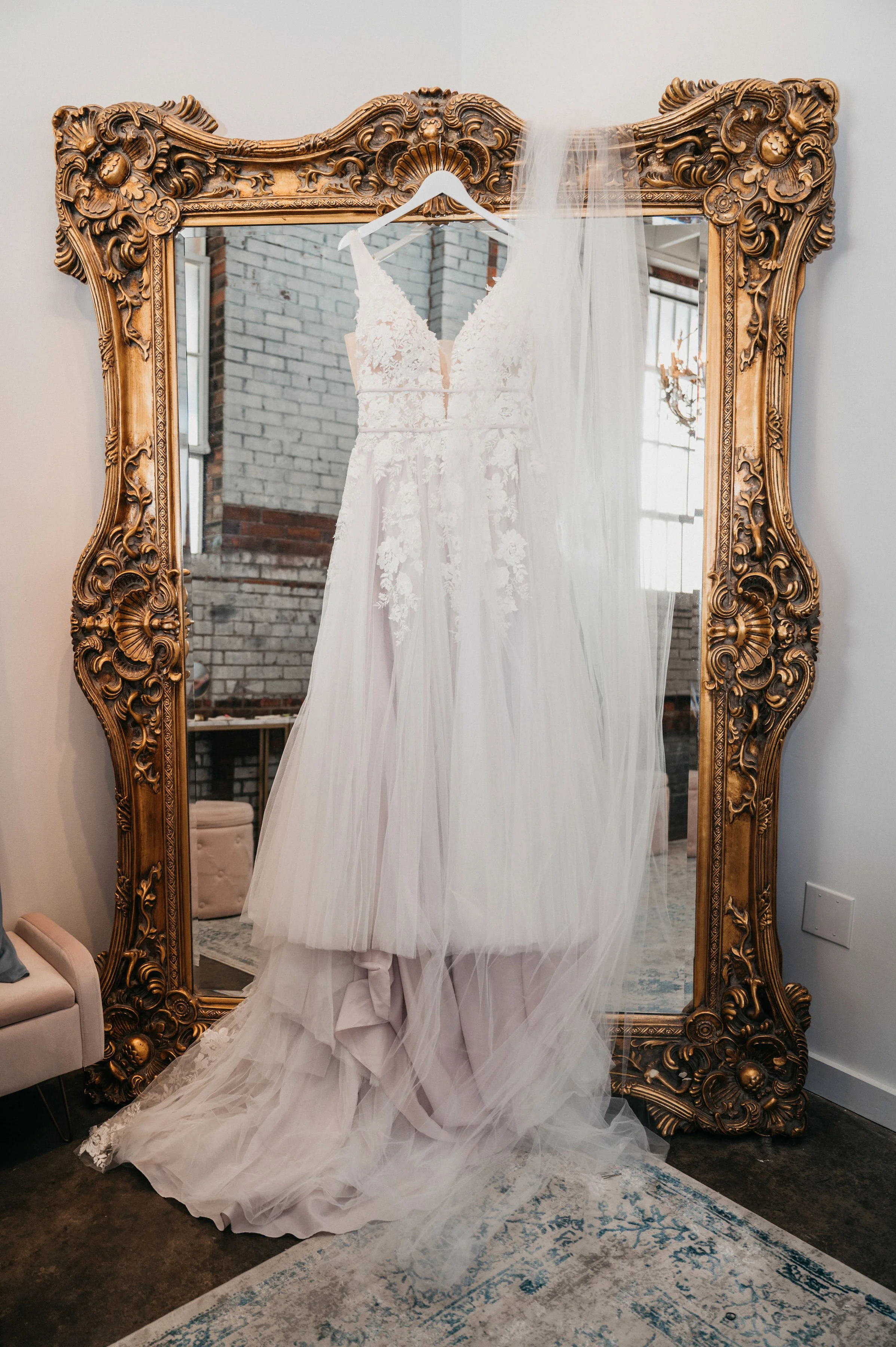 Wedding dress hanging on a white hanger in front of an ornate gold mirror, with a veil draped over the mirror's corner.