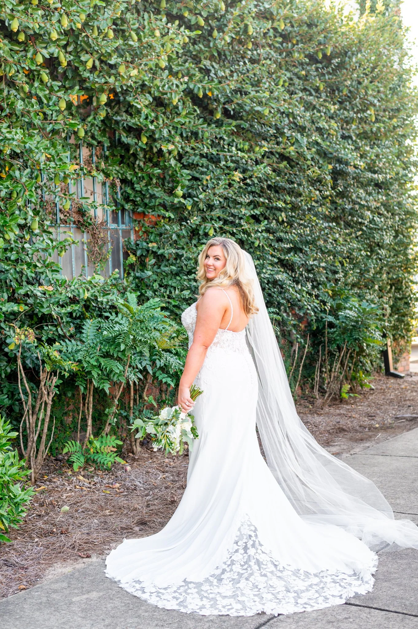 A bride in a white wedding gown with lace details, holding a bouquet, standing on a sidewalk beside a green hedge with small pears, smiling at the camera.
