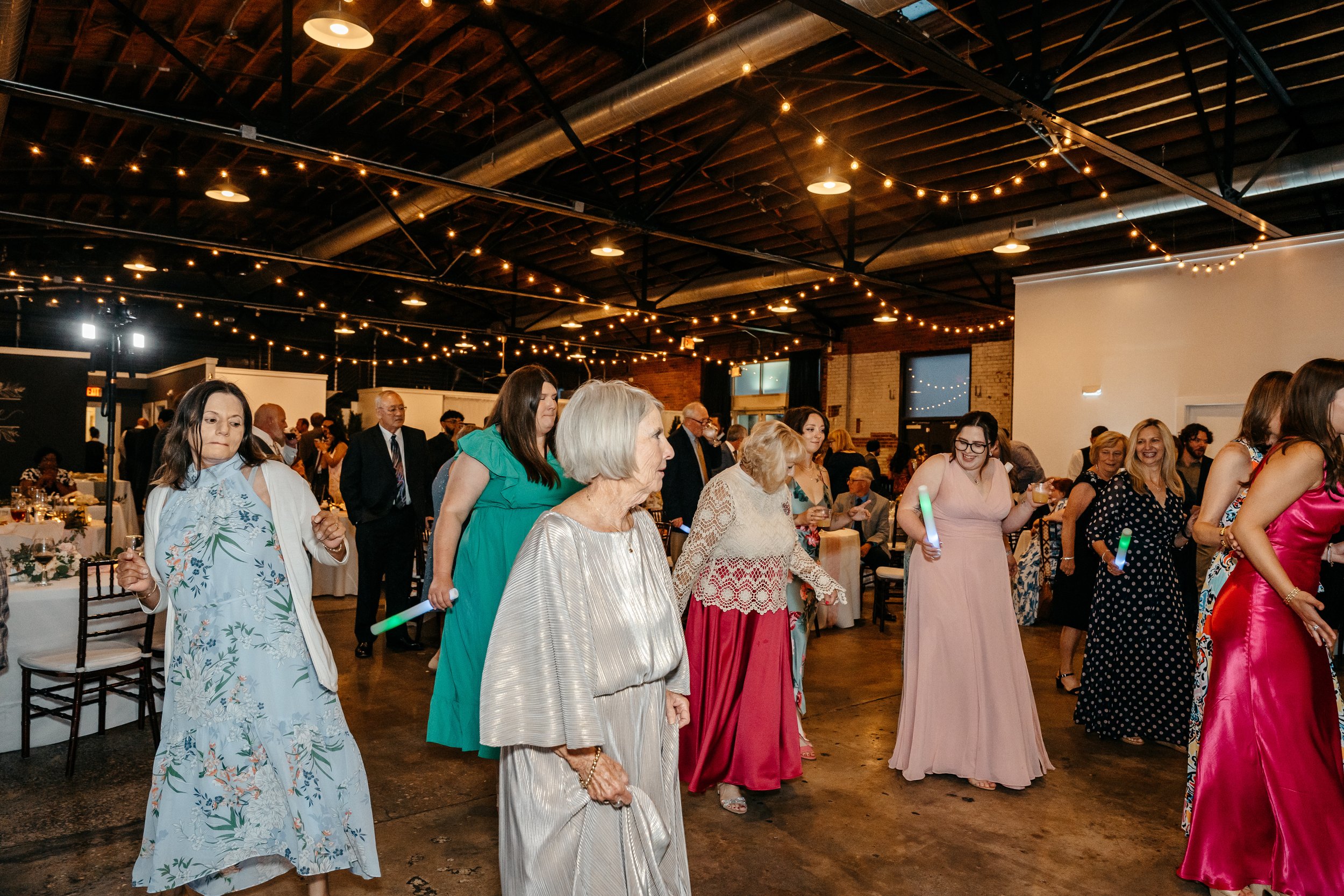 People dancing at an indoor celebration with string lights, varying in ages, dressed in colorful dresses and suits, some holding glow sticks.
