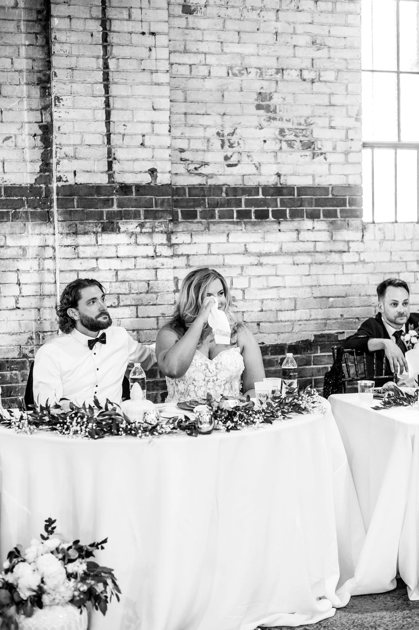 Black and white photo of a bride and groom sitting at a decorated head table during a wedding reception, with a brick wall and window in the background.