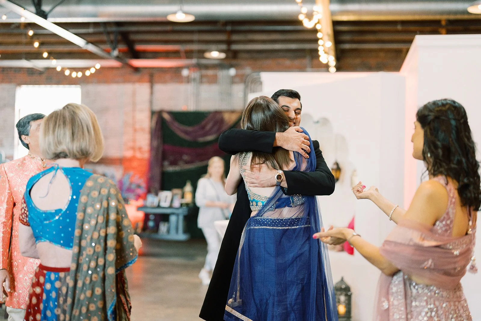 A man in a black suit hugging a woman in a blue sari, surrounded by several women in colorful traditional Indian clothing at a celebration or party in a decorated indoor venue.