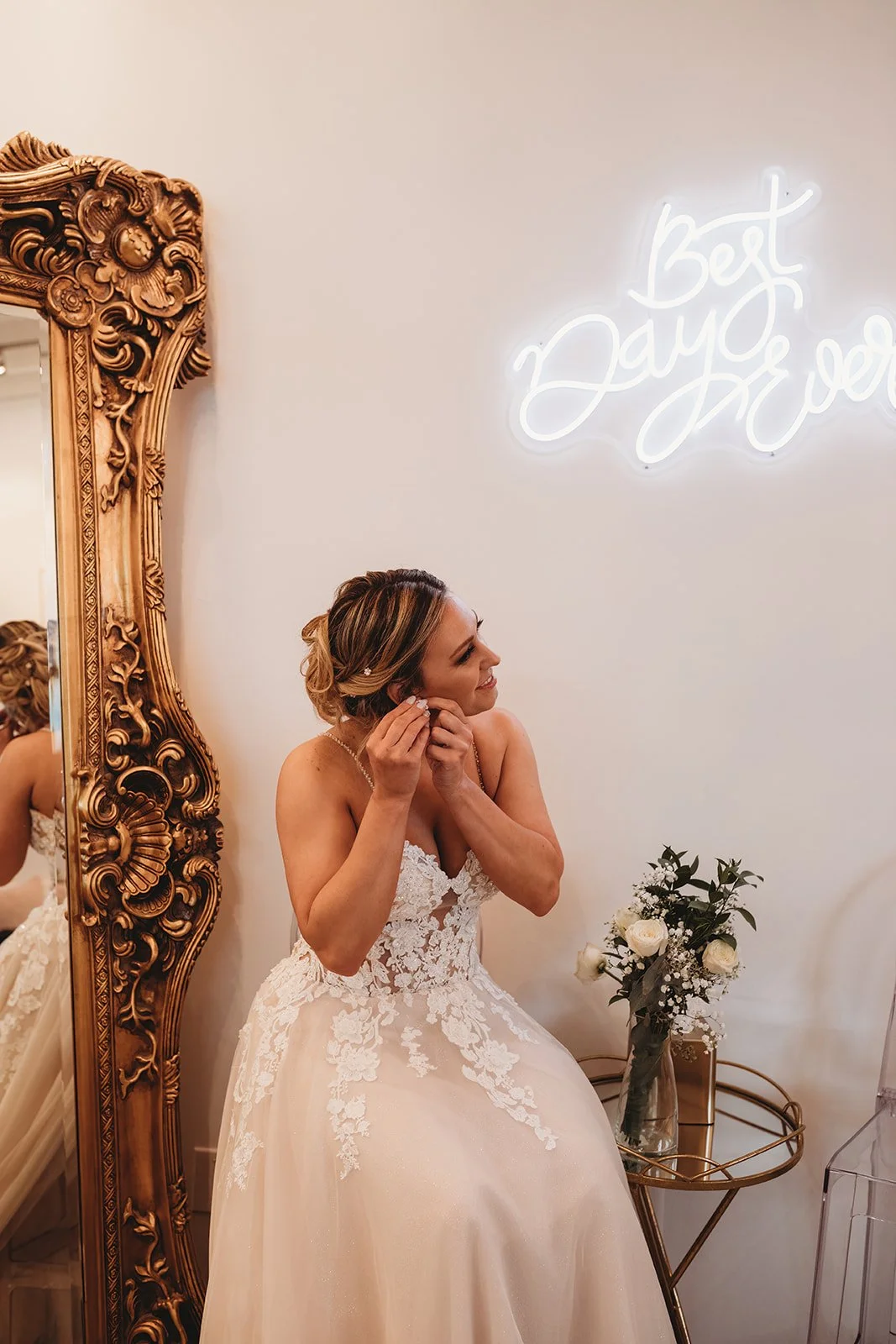 A woman in a wedding dress putting on earrings, smiling, and sitting in front of a large ornate mirror and a vase of white roses. There is a neon sign that says 'Best Day Ever' on the wall behind her.
