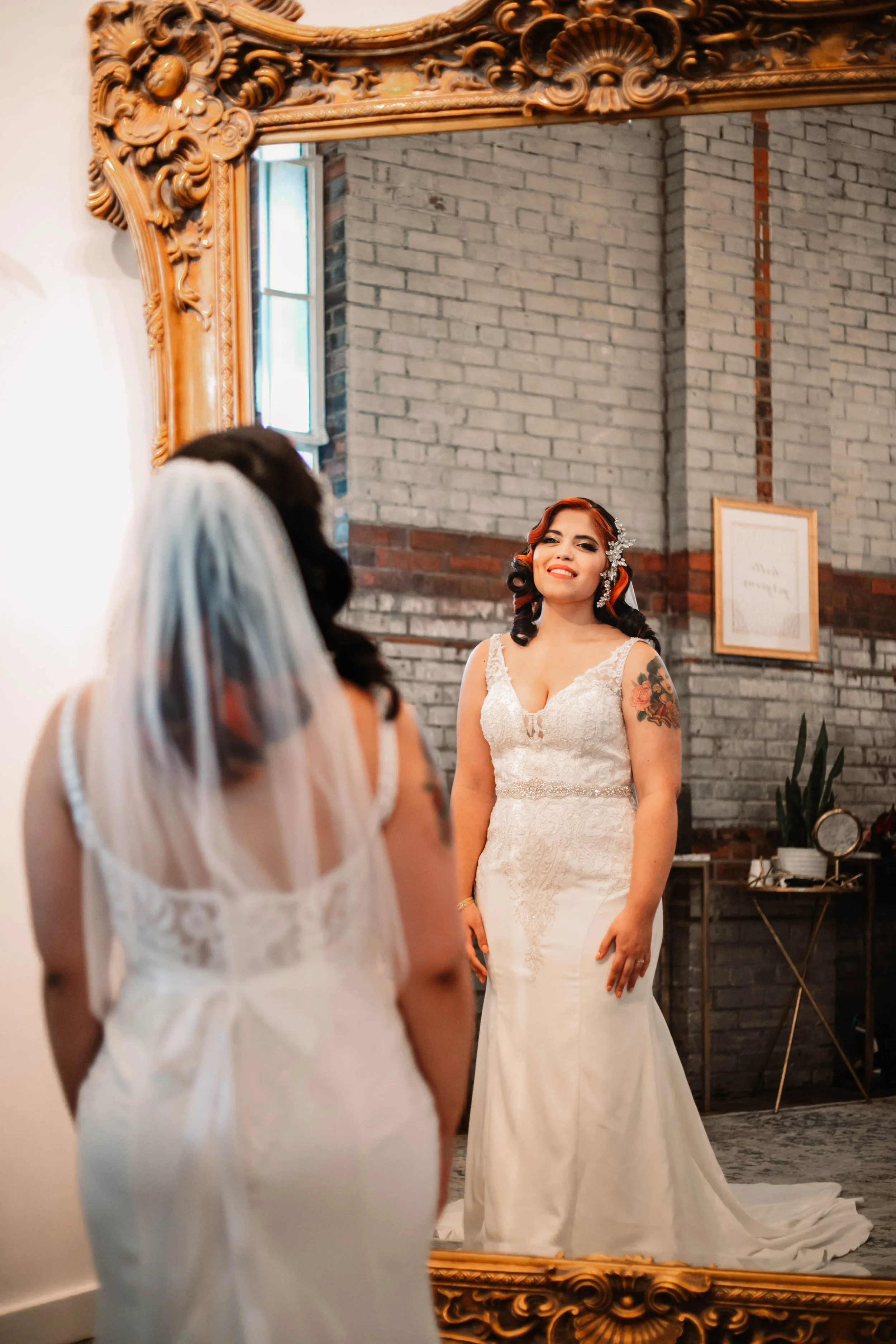 A bride looking at her reflection in a large ornate mirror, wearing a white wedding gown with lace details and a veil, in a room with gray brick walls and a window.